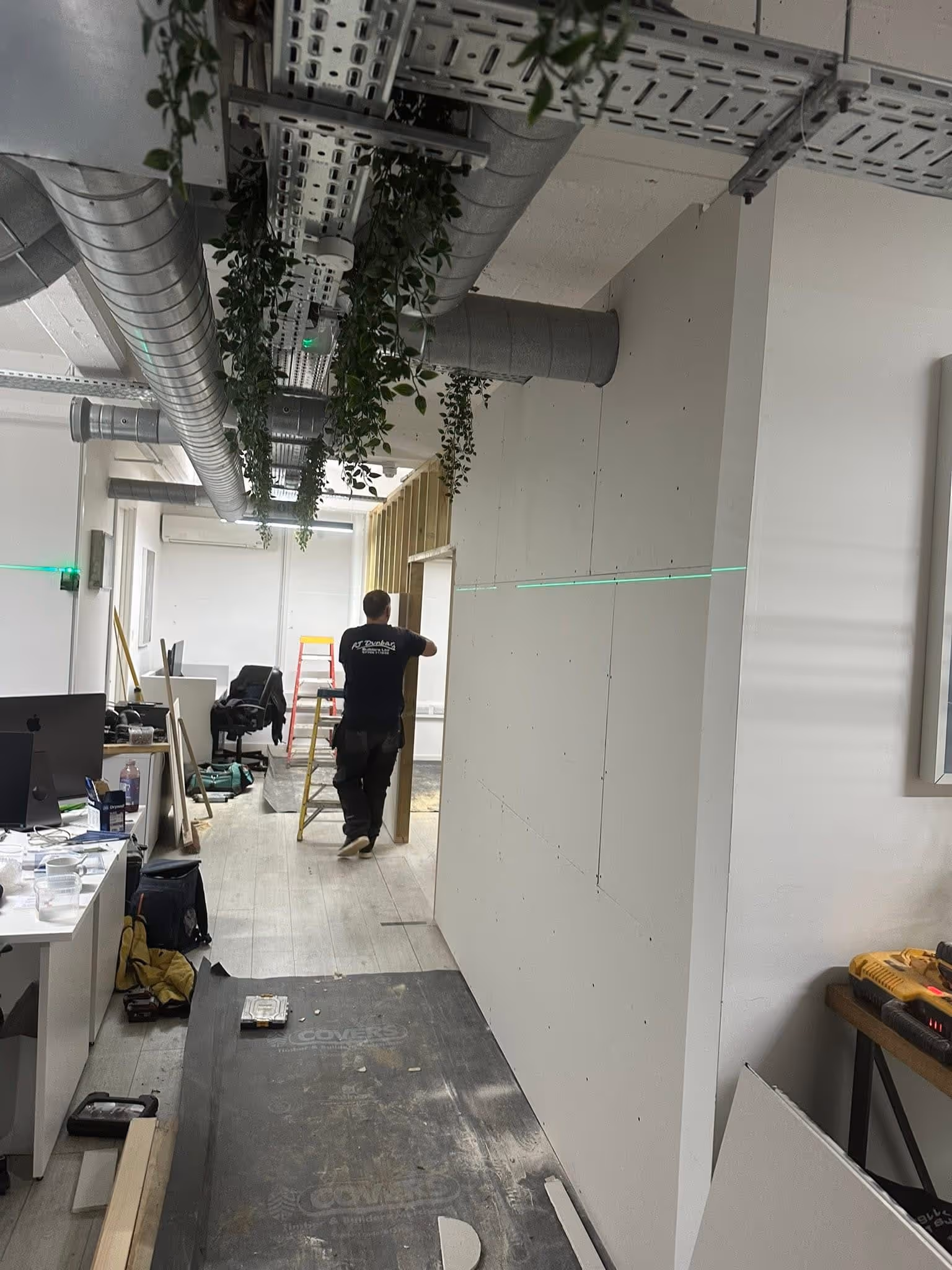 Worker transporting a wooden plank in an office under renovation with exposed ceiling ducts and hanging plants.