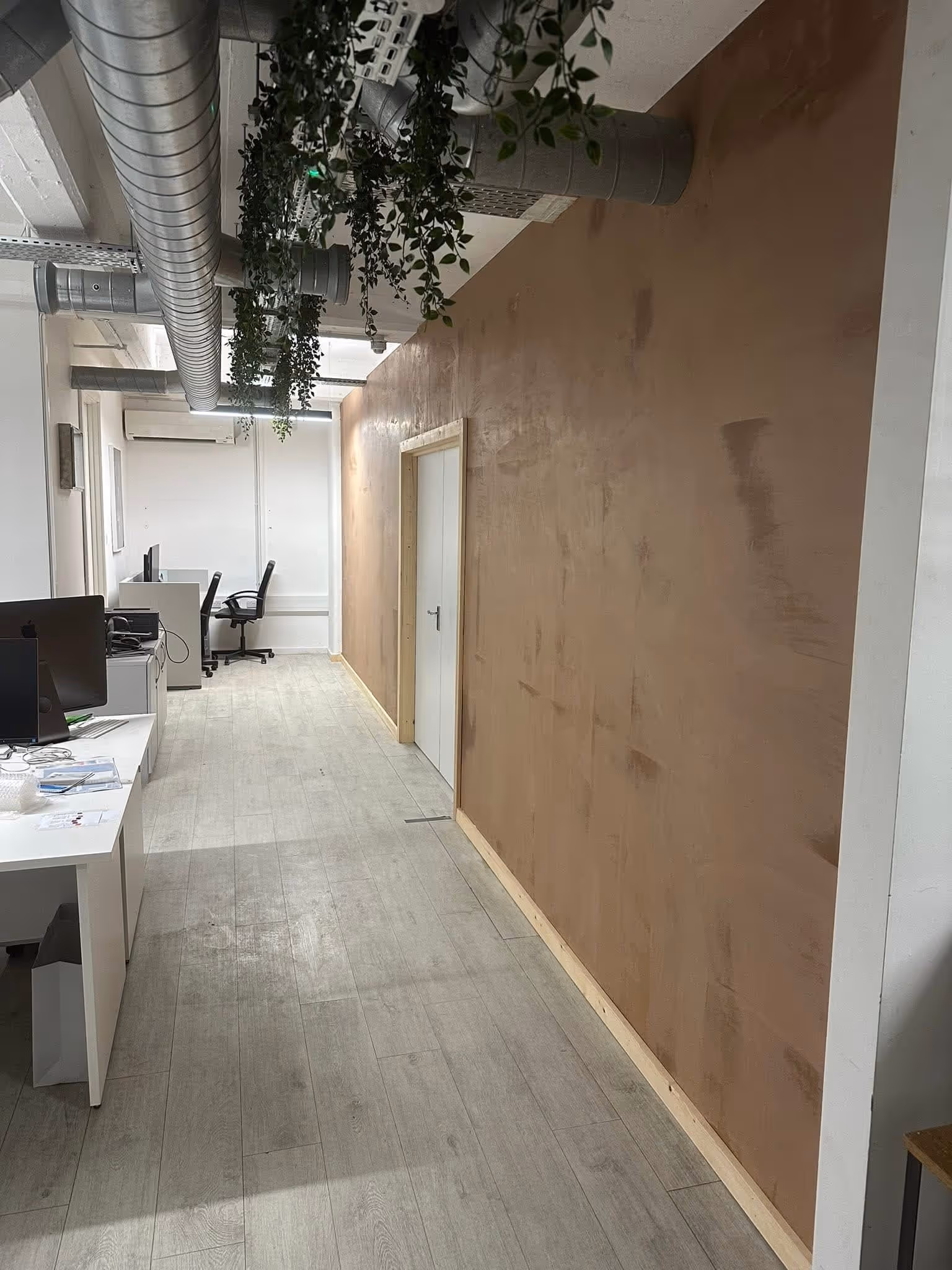 Modern office corridor with light wood flooring, beige plastered wall, white door, white desks with computers, black office chairs, and overhead ventilation pipes with hanging plants.