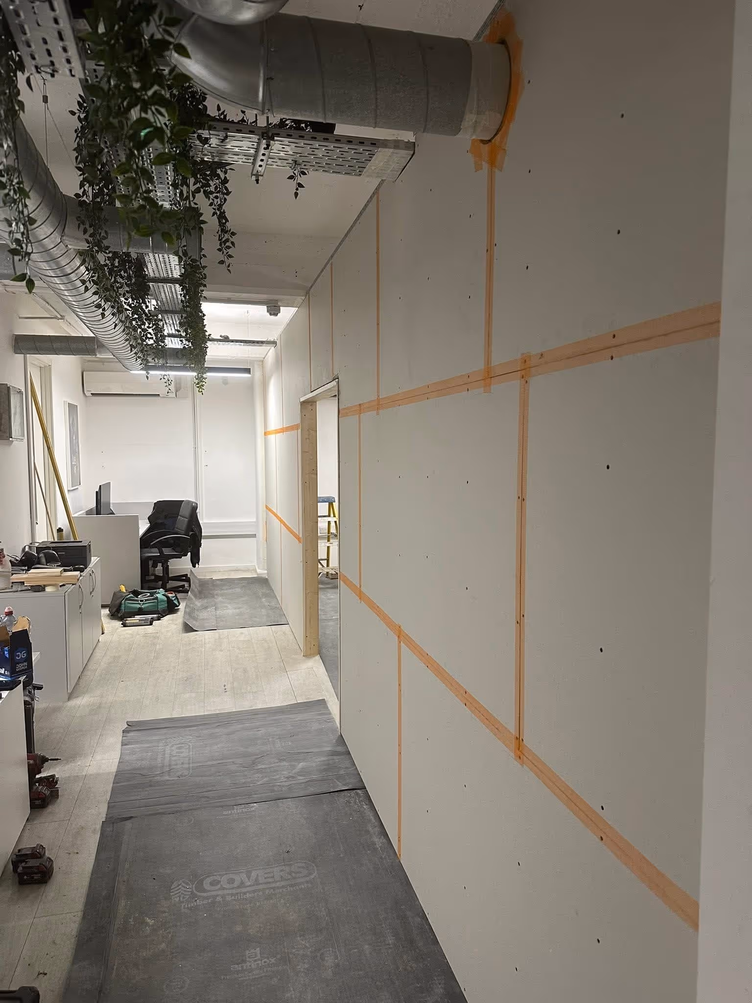 Office corridor under renovation with protective floor mats, drywall walls with orange tape seams, overhead ventilation ducts, and some office furniture.