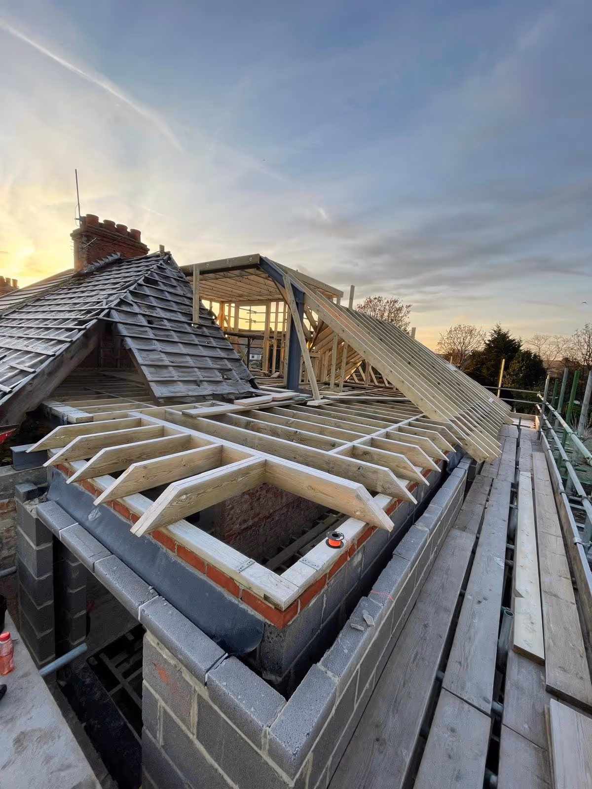 Partially constructed roof extension with wooden framing and scaffolding at sunset.