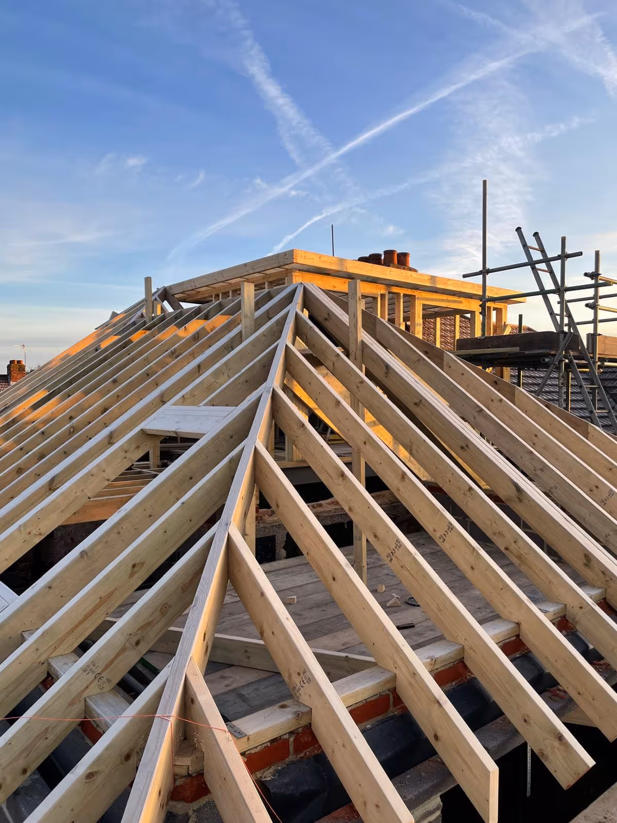 Wooden roof frame structure under construction with clear sky and scaffolding in the background.