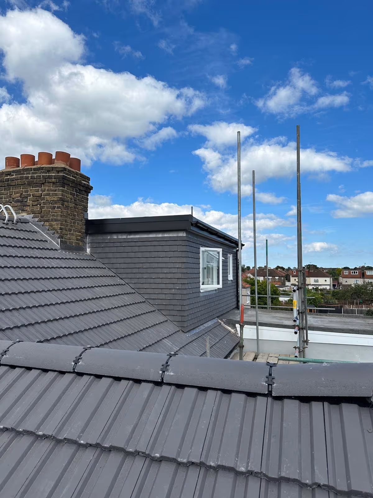 Gray tiled rooftops with a brick chimney, a small windowed dormer, and metal scaffolding under a partly cloudy blue sky.