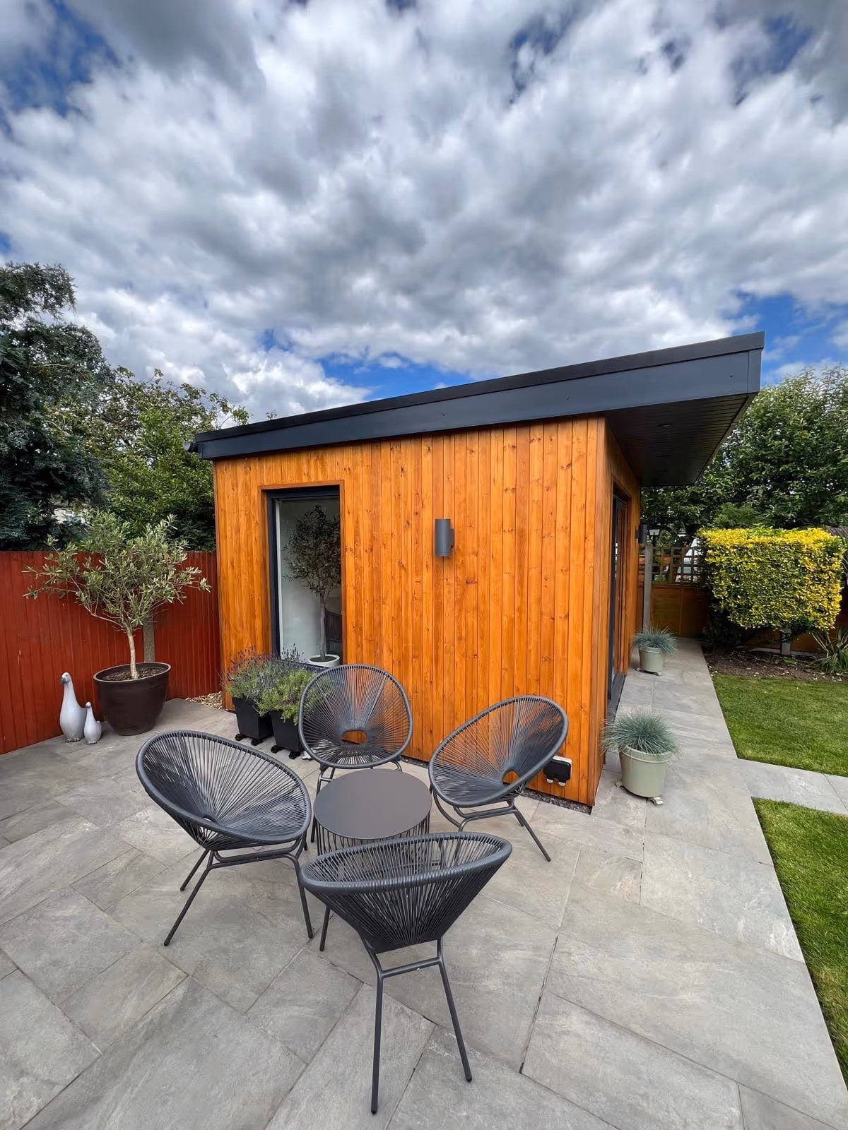 Modern wooden garden room with slanting roof and patio area featuring four black woven chairs and a small round table.