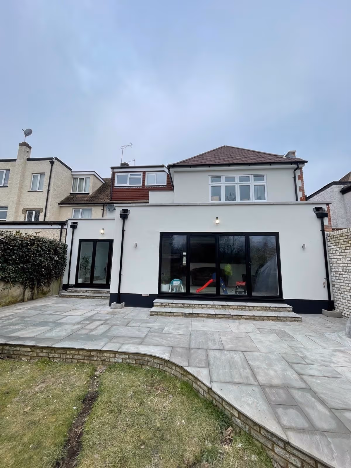 Rear view of a modern two-story house with large glass sliding doors opening to a tiled patio and a lawn in the foreground.