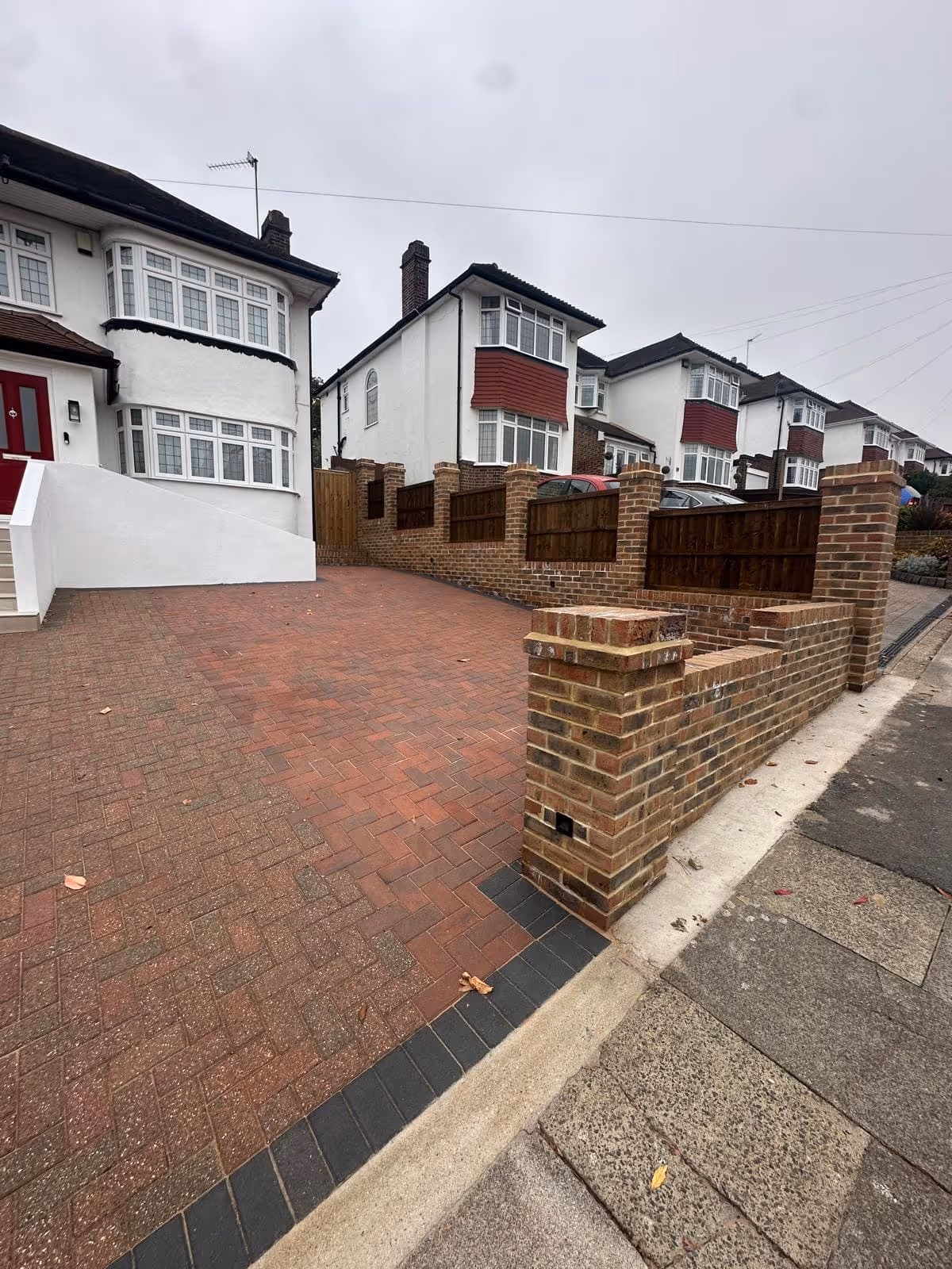 Brick-paved driveway with low brick wall and wooden fence panels, leading to white houses with bay windows on a cloudy day.