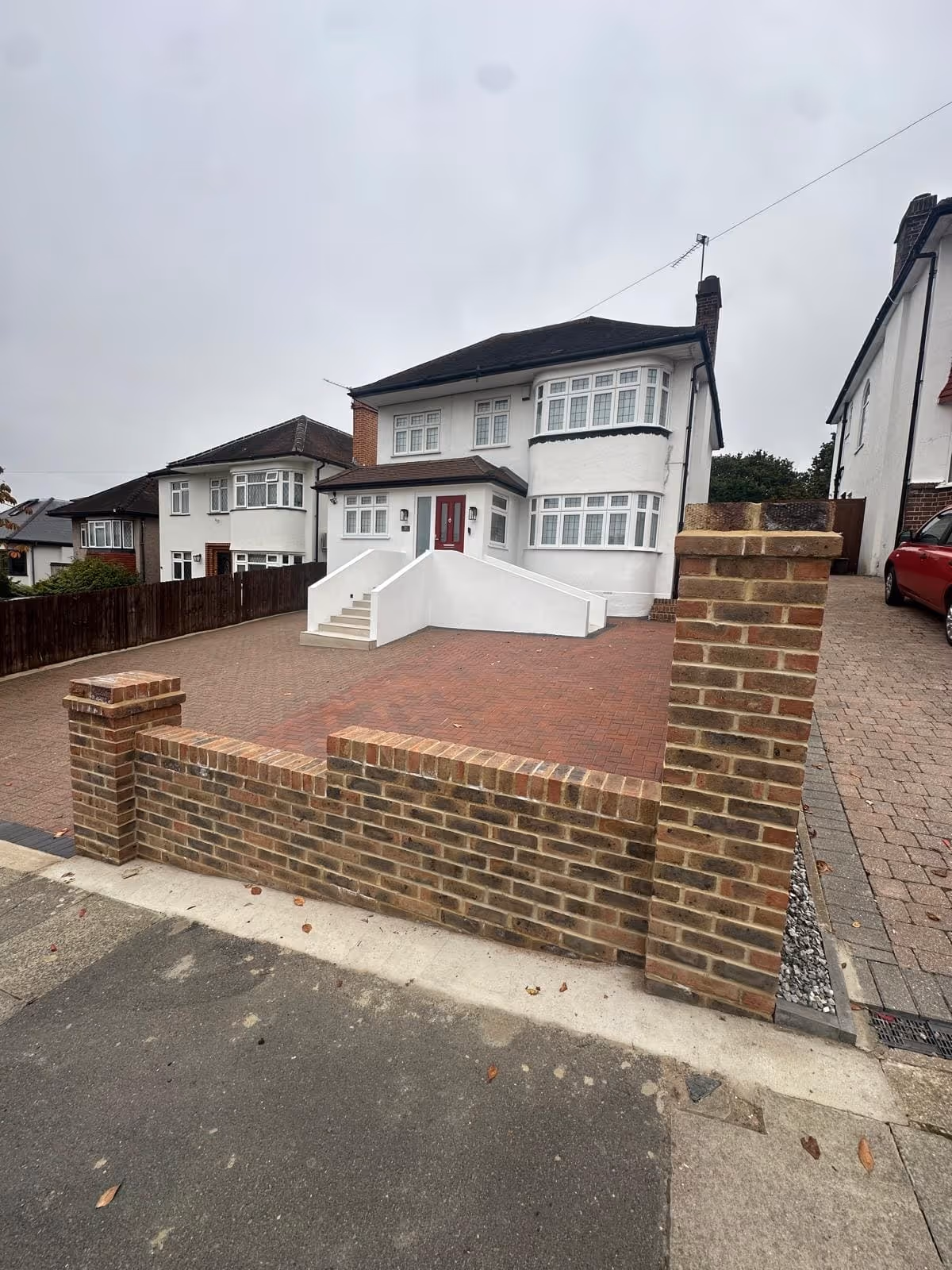 Two-story white house with bay windows, a red door, front steps with white railings, a paved driveway, and a low brick wall bordering the sidewalk.