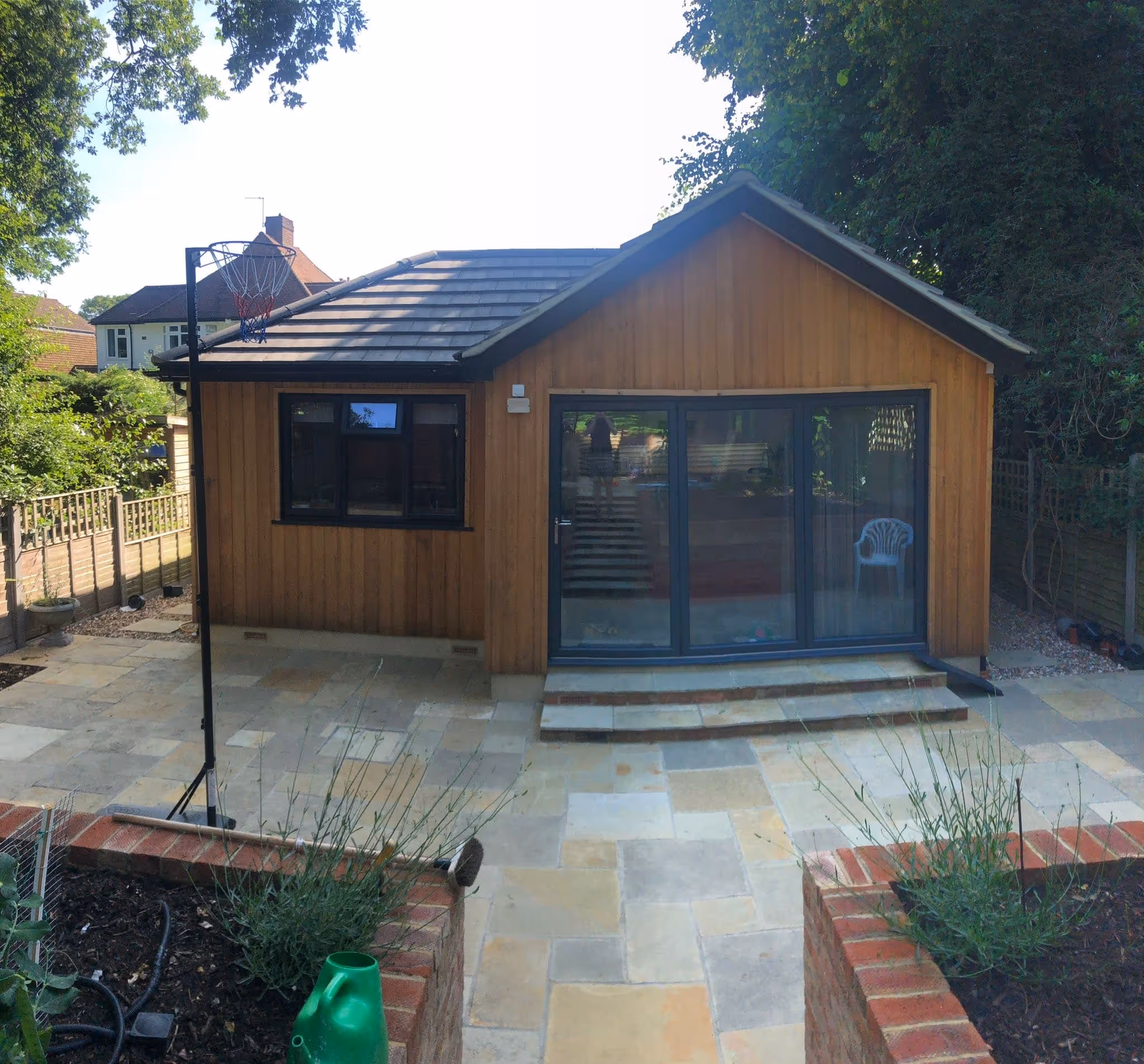 Small wooden garden house with black-framed window and glass sliding doors opening onto a paved patio with a basketball hoop on the left.