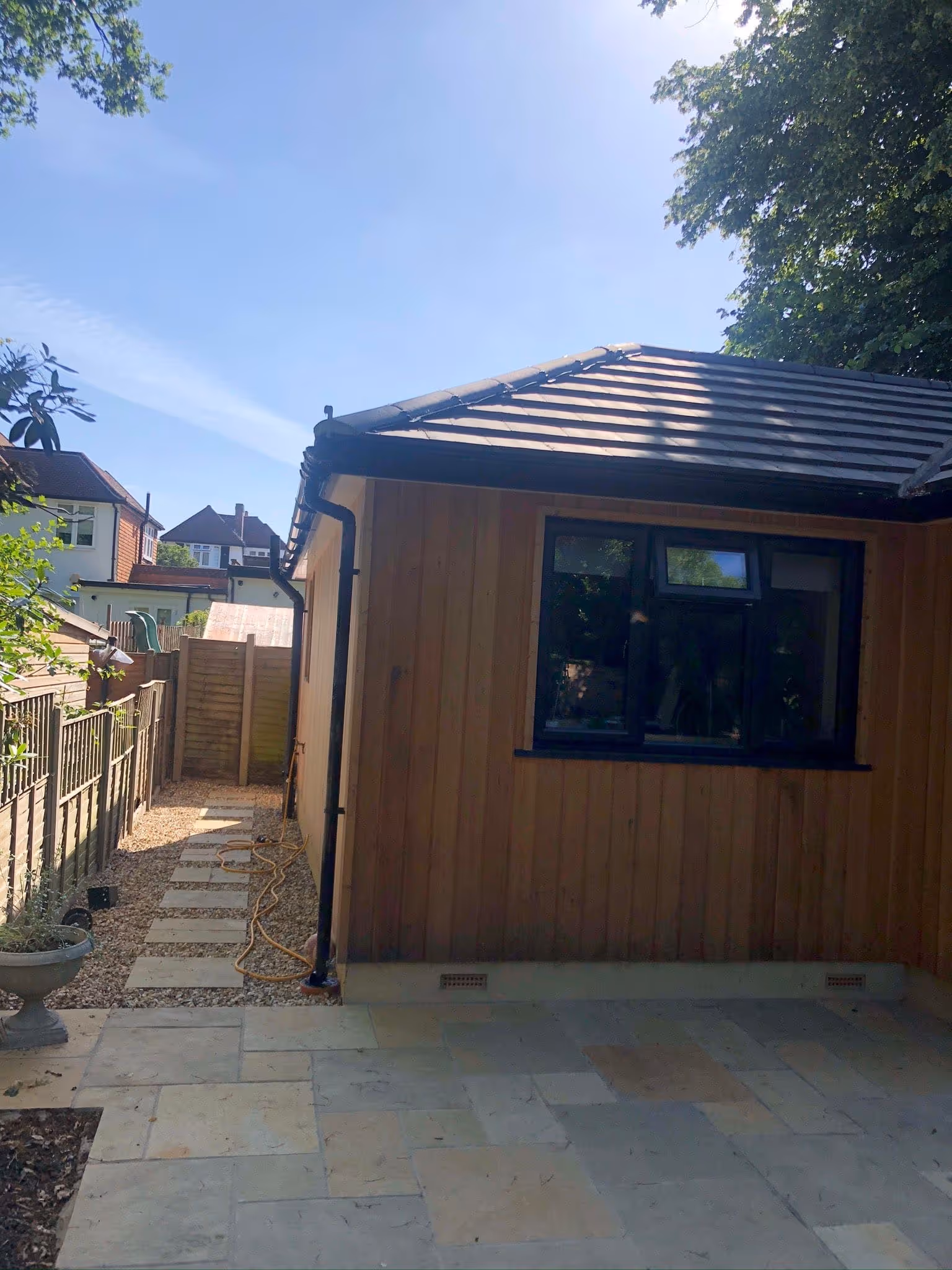 Backyard with stone patio, wooden shed with black window frames, and a gravel path alongside a fenced garden under a clear blue sky.
