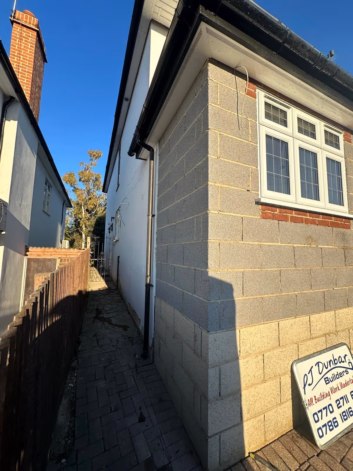 Side view of a house under construction with brick and block walls, a narrow paved pathway, and a sign for PJ Dunbar Builders leaning against the wall.