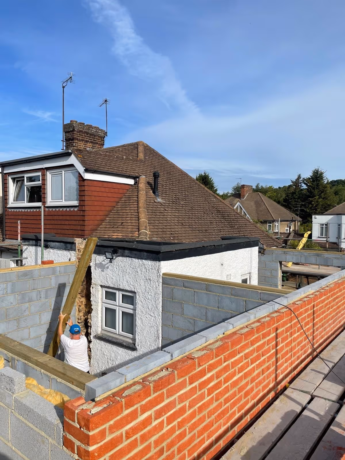 Construction worker installing wooden beam inside partially built brick and cinder block walls next to a house with a brown roof under a blue sky.