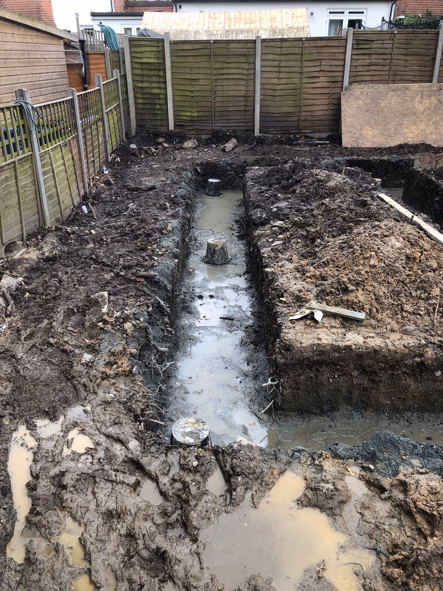 Construction site with muddy ground and trenches excavated for foundation work, surrounded by wooden fences.