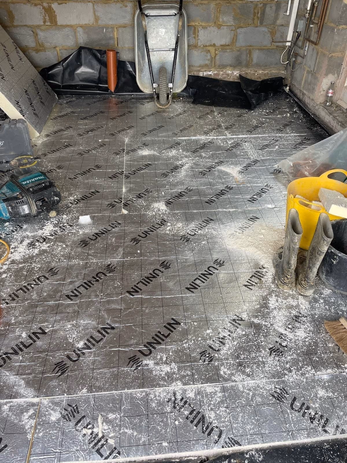 Construction site floor covered with insulation panels labeled UNILIN, with a wheelbarrow, muddy boots, yellow bucket, and power tools.