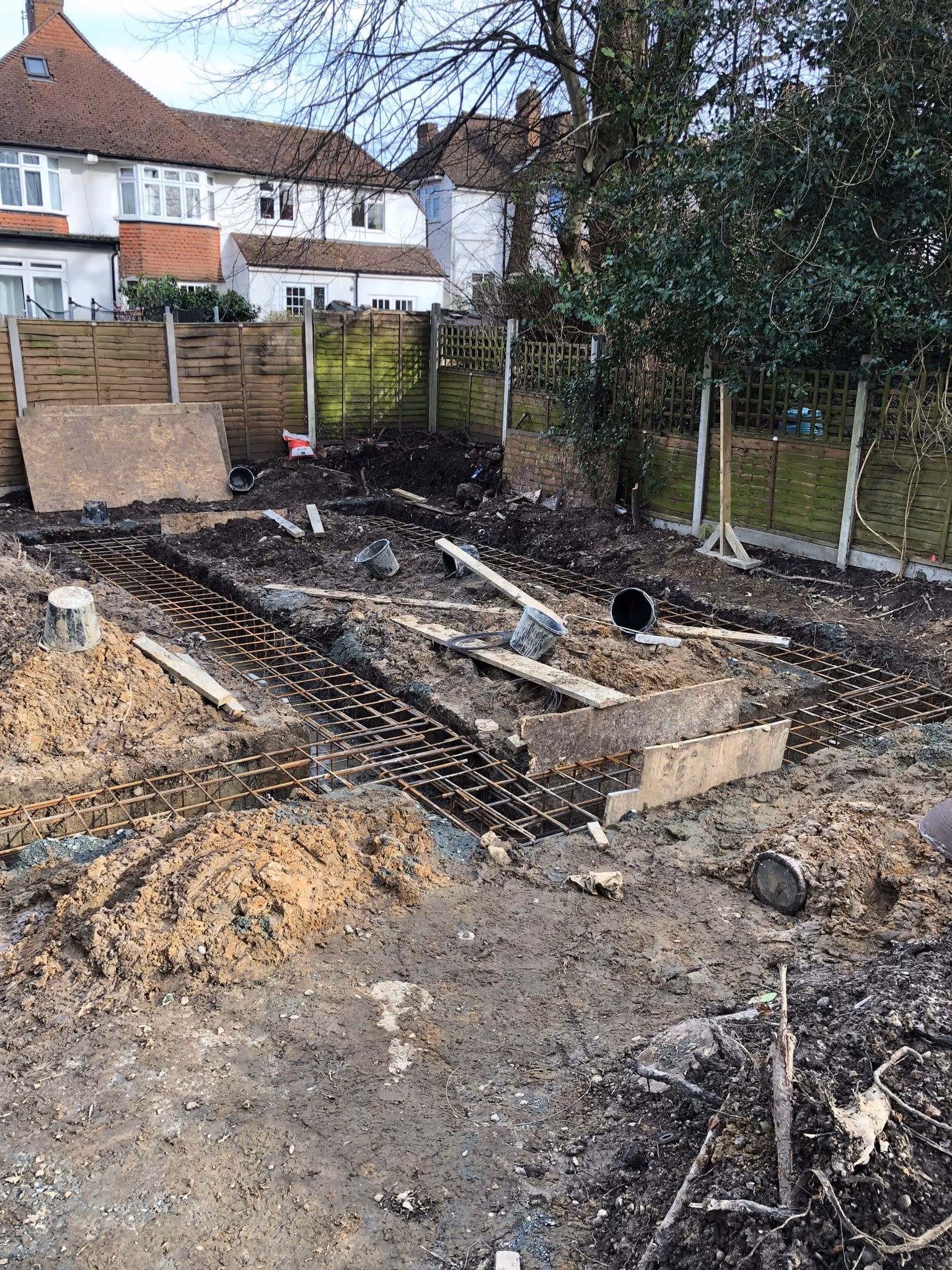 Construction site in a backyard with exposed rebar grids for a foundation and piles of dirt surrounded by wooden fences and residential houses.