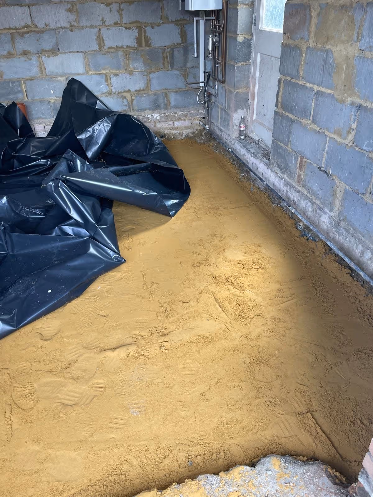 Indoor construction area with compacted sandy soil floor, black plastic sheet partially covering one side, and unfinished cinder block walls.