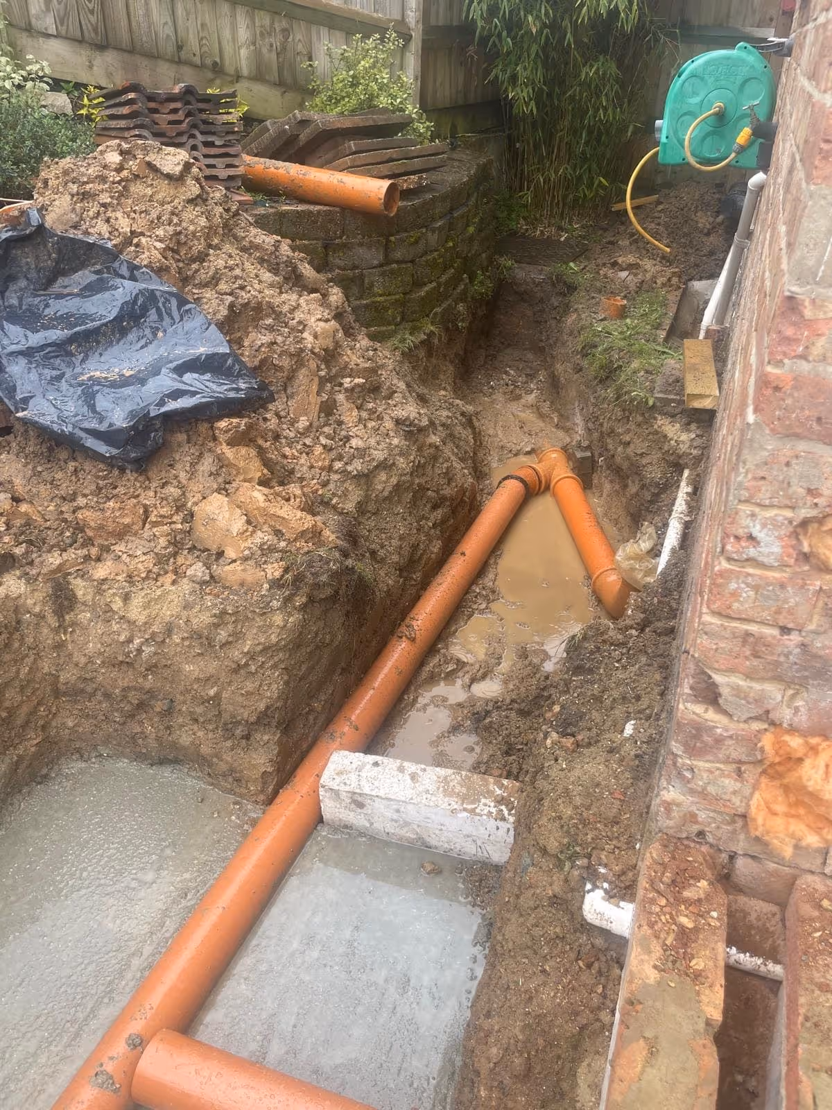 Construction site with partially installed orange drainage pipes in a muddy trench next to a brick wall.