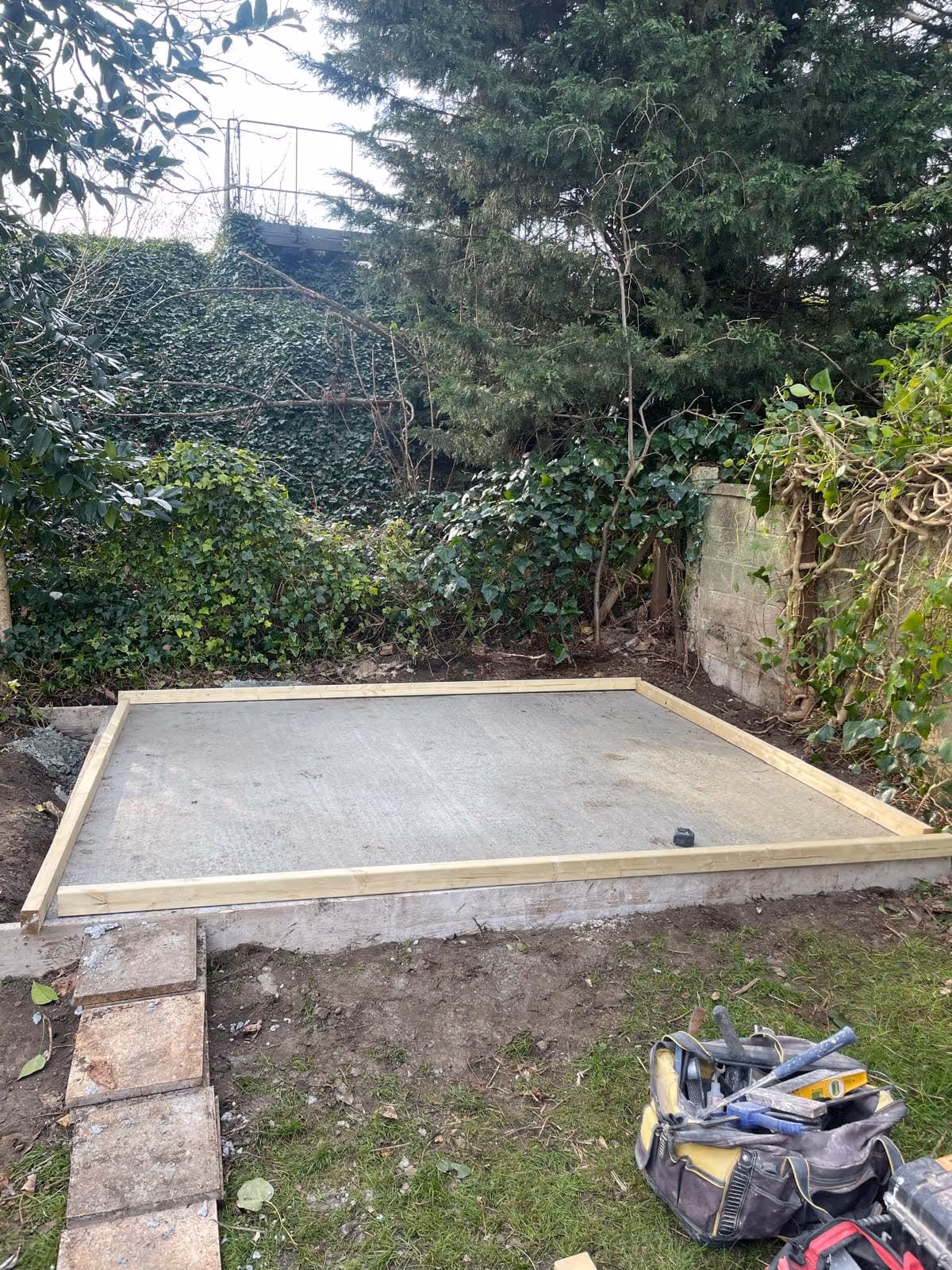 Concrete foundation slab with wooden framing in a garden surrounded by greenery, with a tool bag and construction tools nearby.