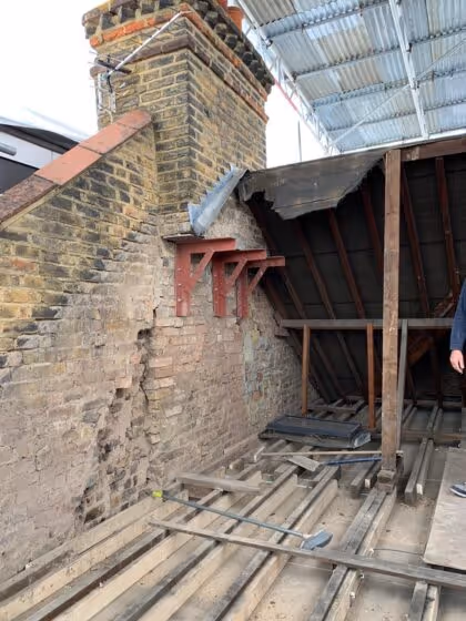 Partially demolished attic space with exposed brick chimney, wooden floor joists, and metal chimney support brackets installed on the chimney.