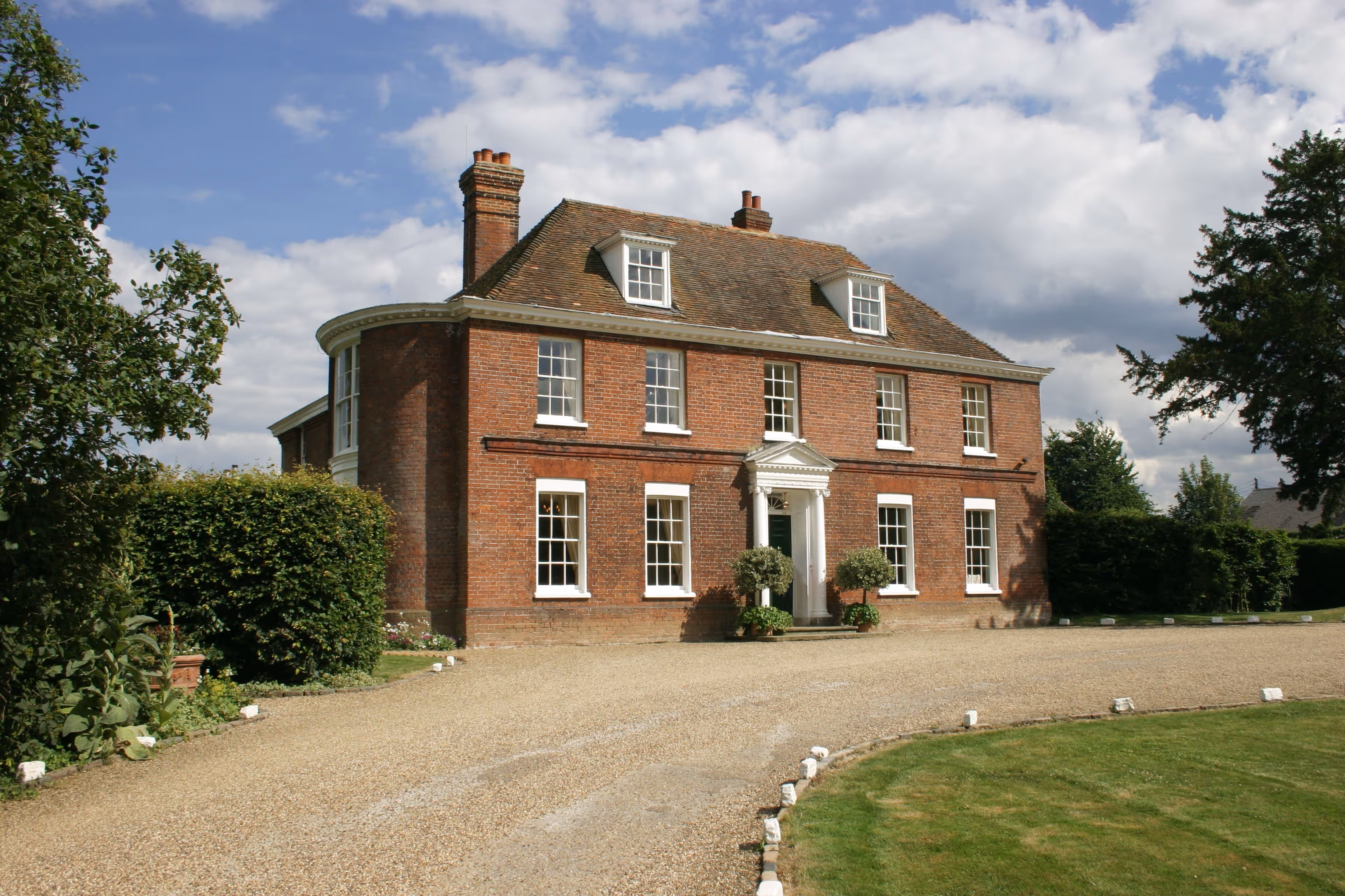 Large traditional red brick house with white-trimmed windows, a gravel driveway, and greenery under a partly cloudy sky.