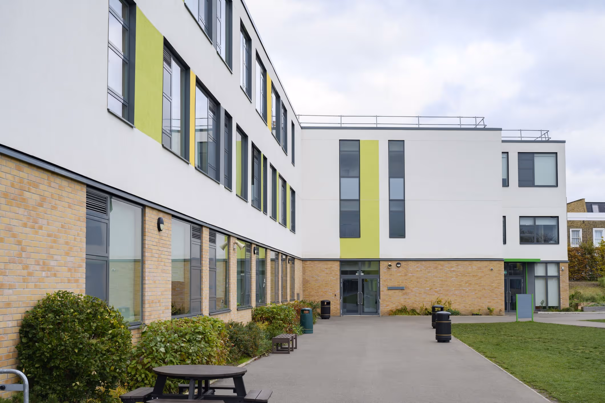 Modern three-story building with white and brick facade, green accent panels, large windows, benches, and trash bins along a paved path.