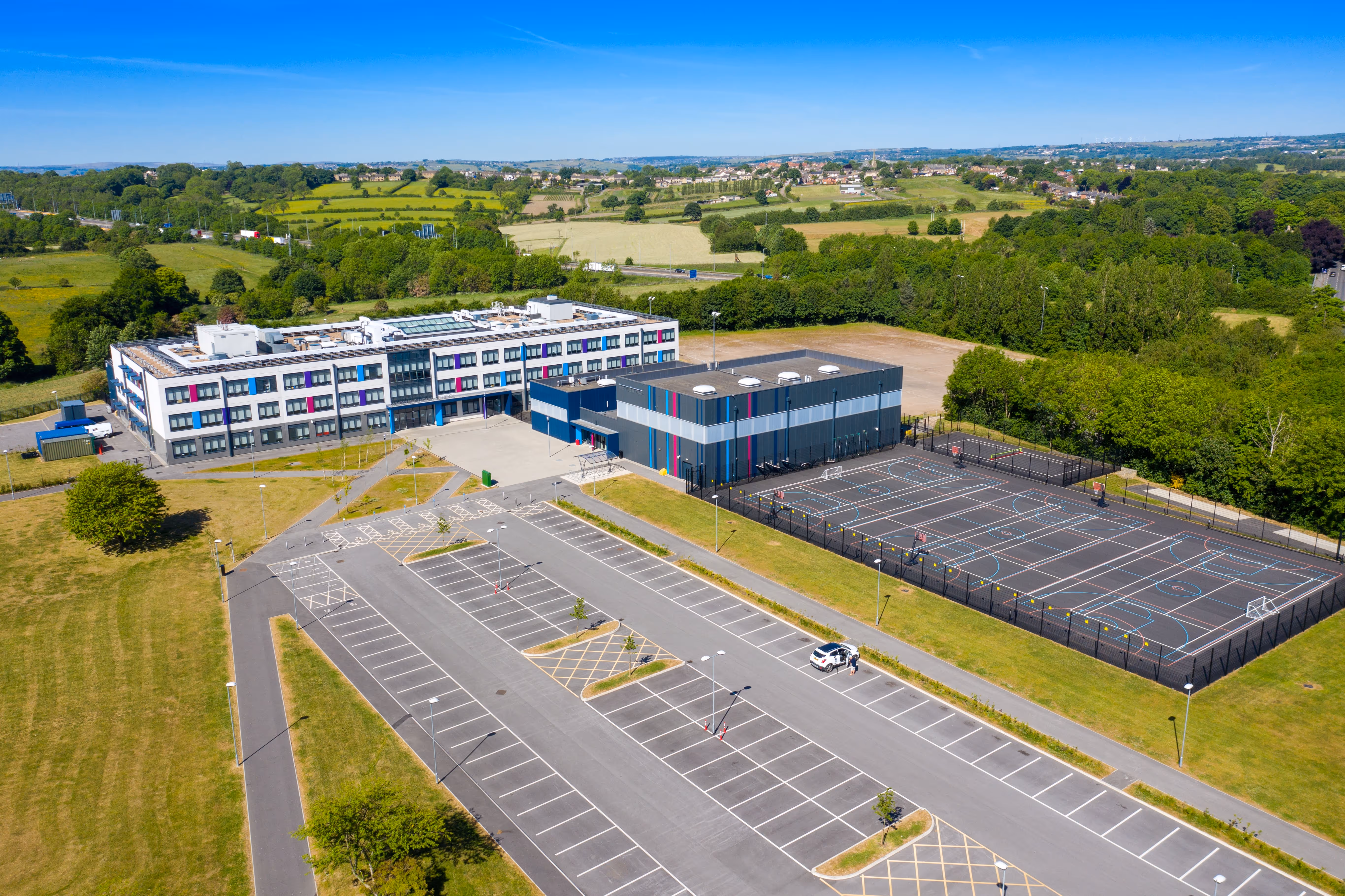 Aerial view of a modern school with a large parking lot, multi-sport courts, and surrounding green fields under a clear blue sky.