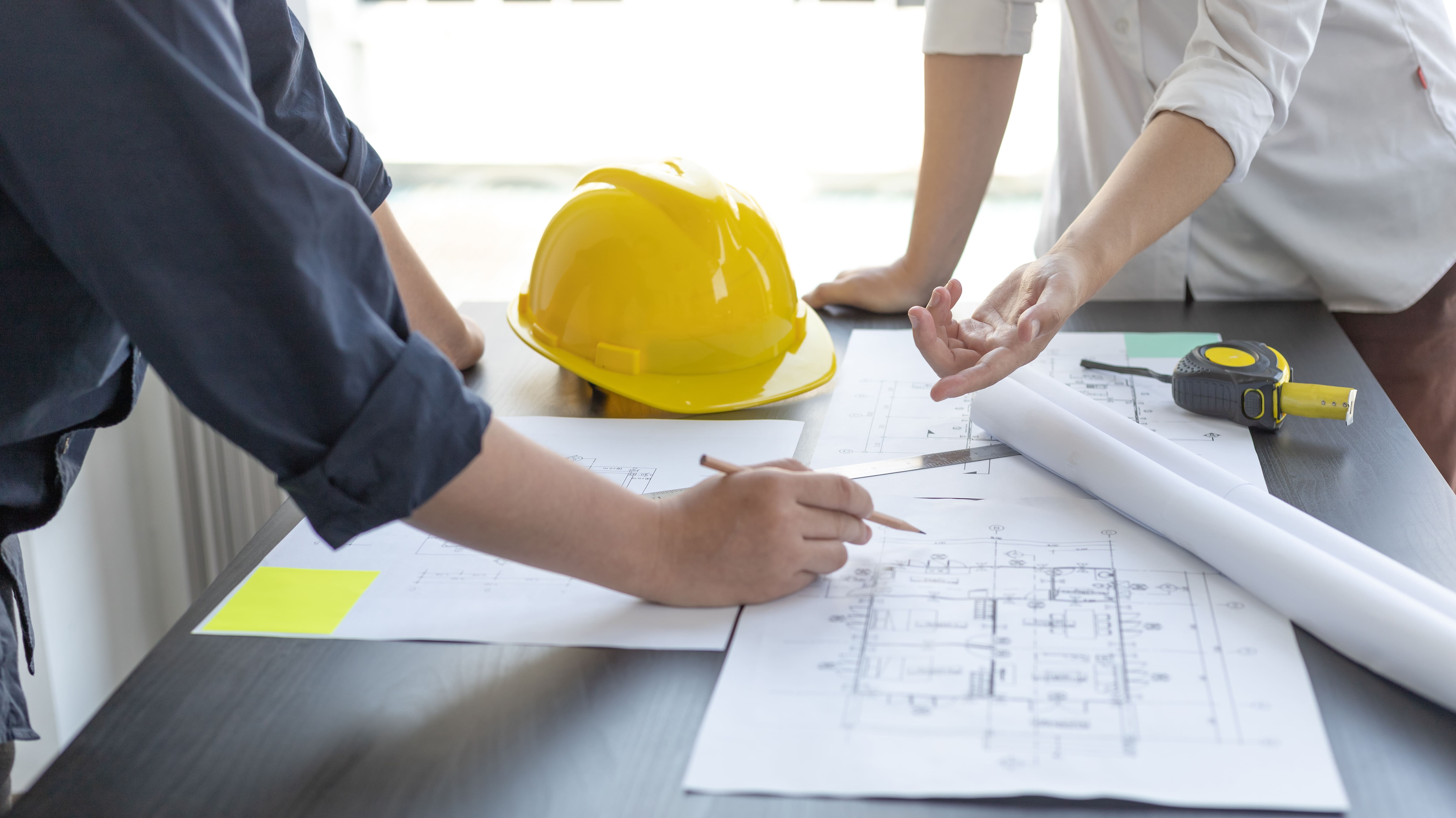 Two people reviewing architectural blueprints on a table with a yellow hard hat and measuring tape.