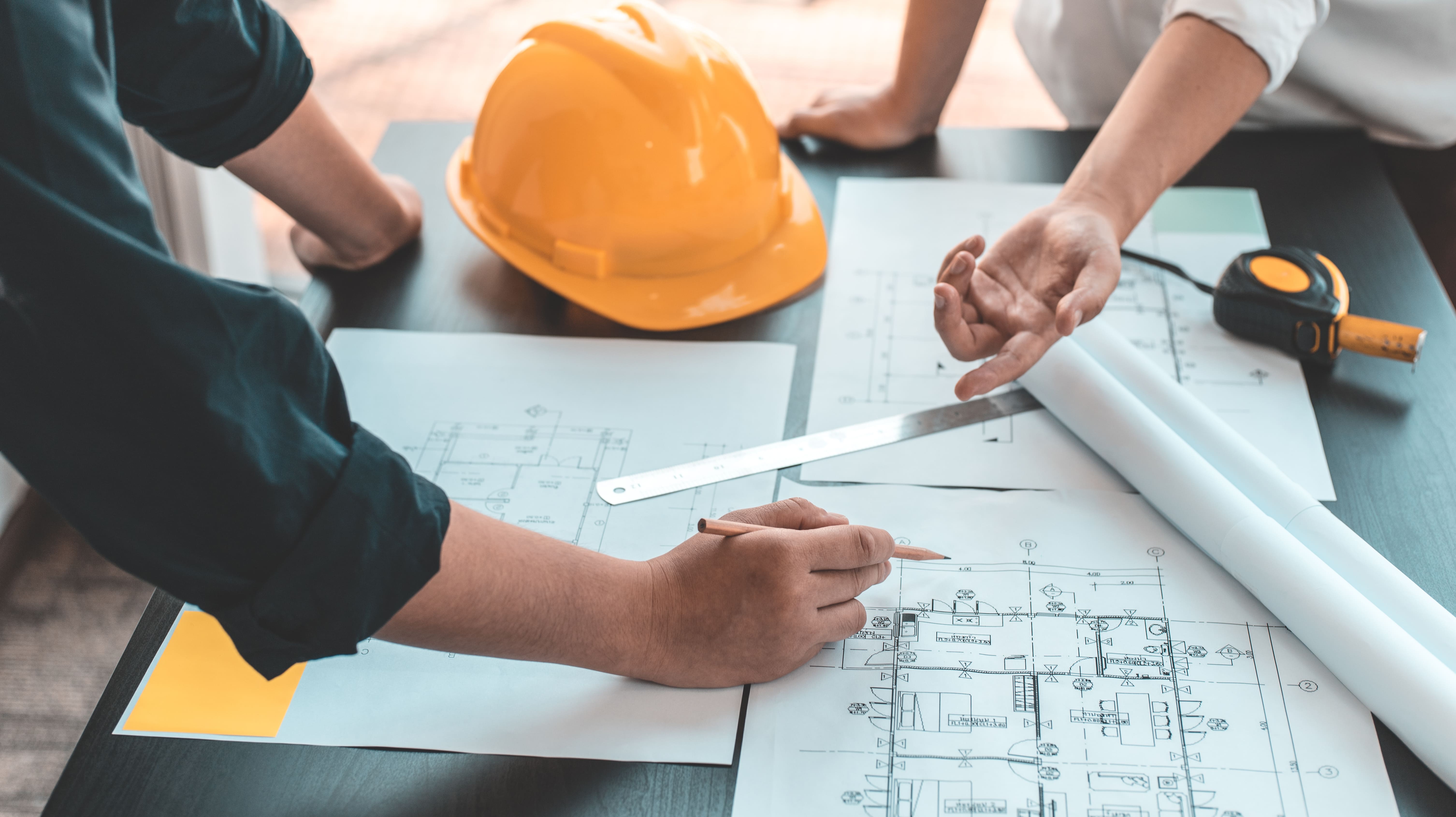 Two people reviewing architectural blueprints on a table with a yellow hard hat and measuring tape.