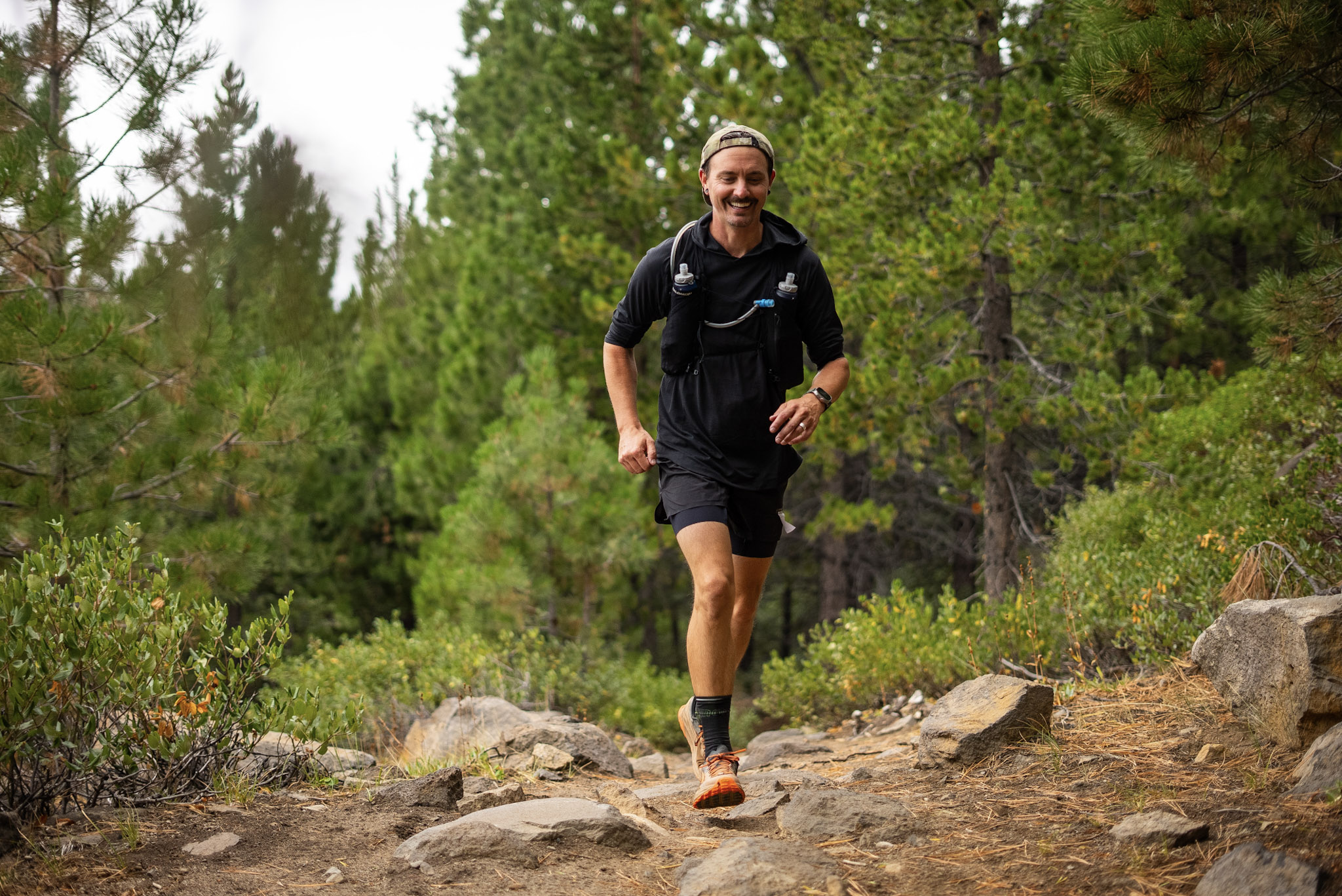 Smiling man wearing a cap and hydration vest trail running on a rocky forest path.