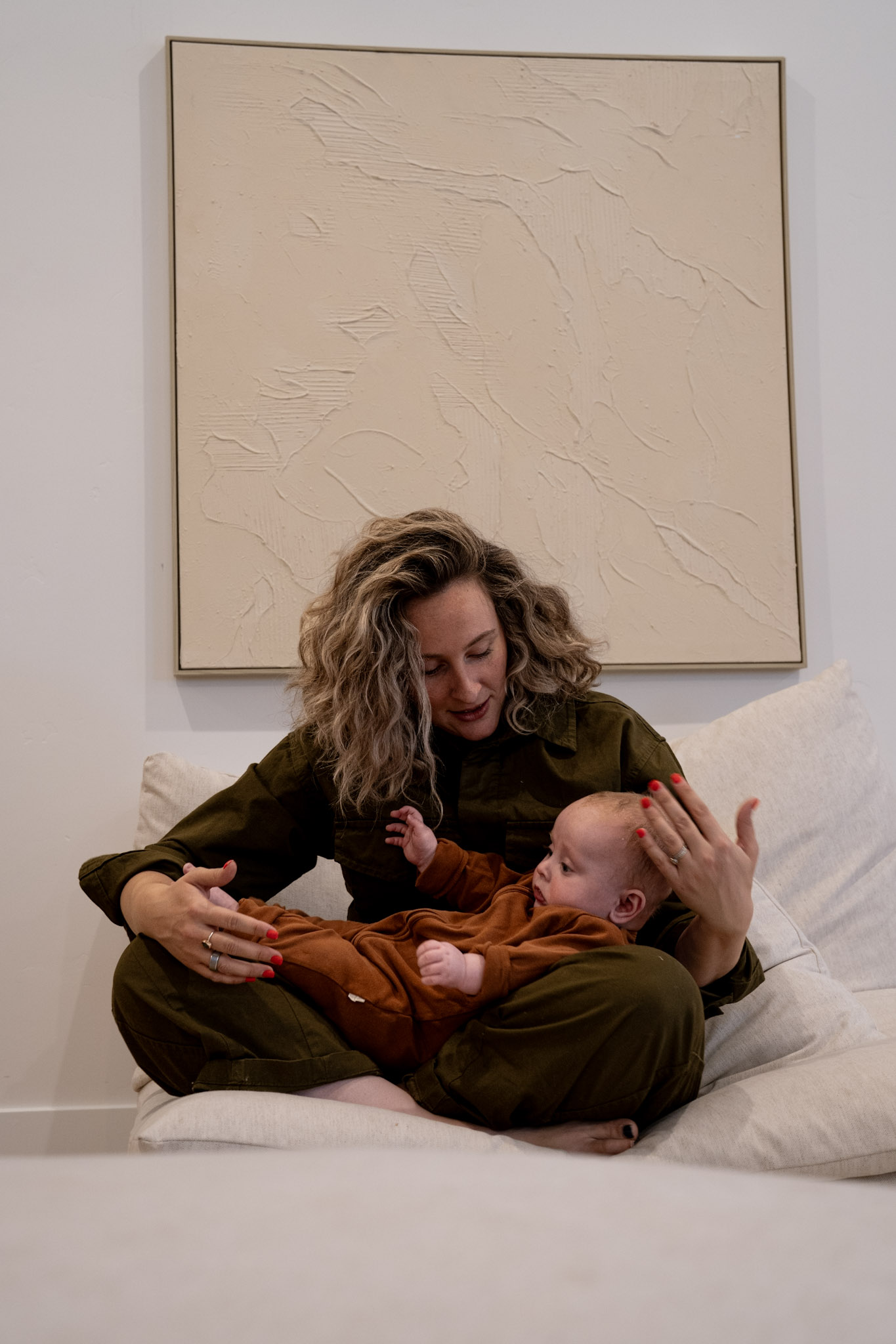 Woman with curly hair sitting cross-legged on a couch holding and looking at a baby dressed in brown.
