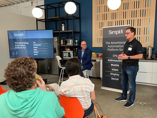 Man in black shirt giving a presentation on an advertising automation platform to an audience in a modern office kitchen.