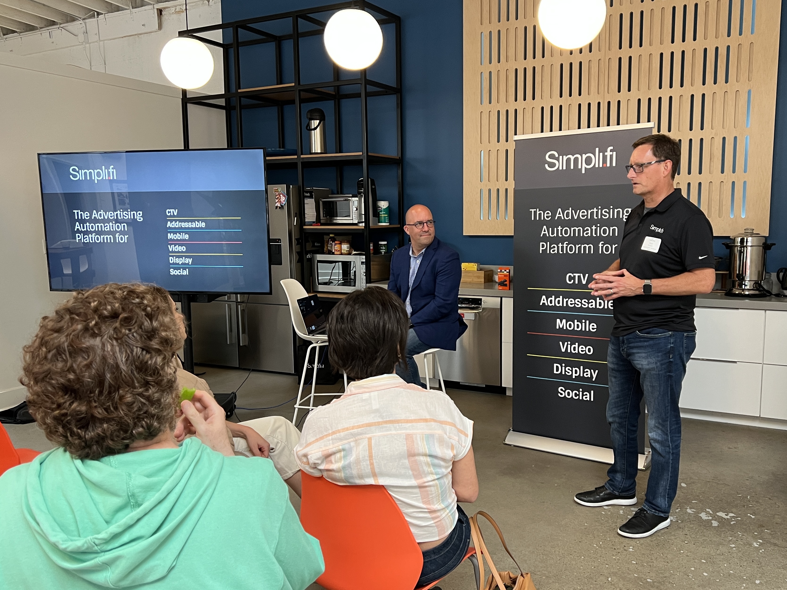 Man in black shirt giving a presentation on an advertising automation platform to an audience in a modern office kitchen.