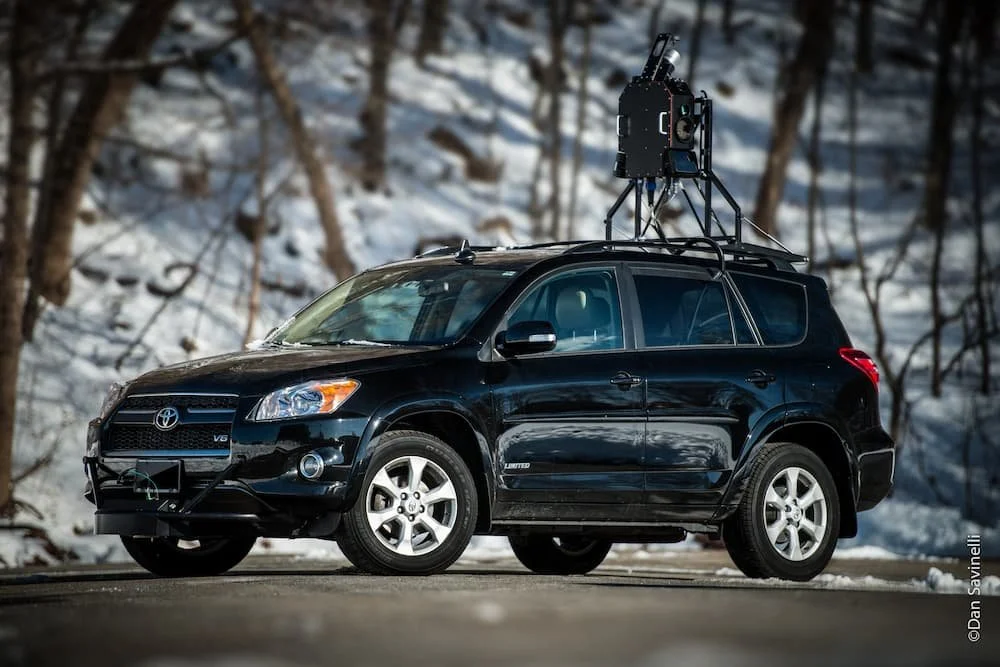 Black Toyota RAV4 SUV with a large mounted camera rig on the roof, parked on a snowy road with trees in the background.