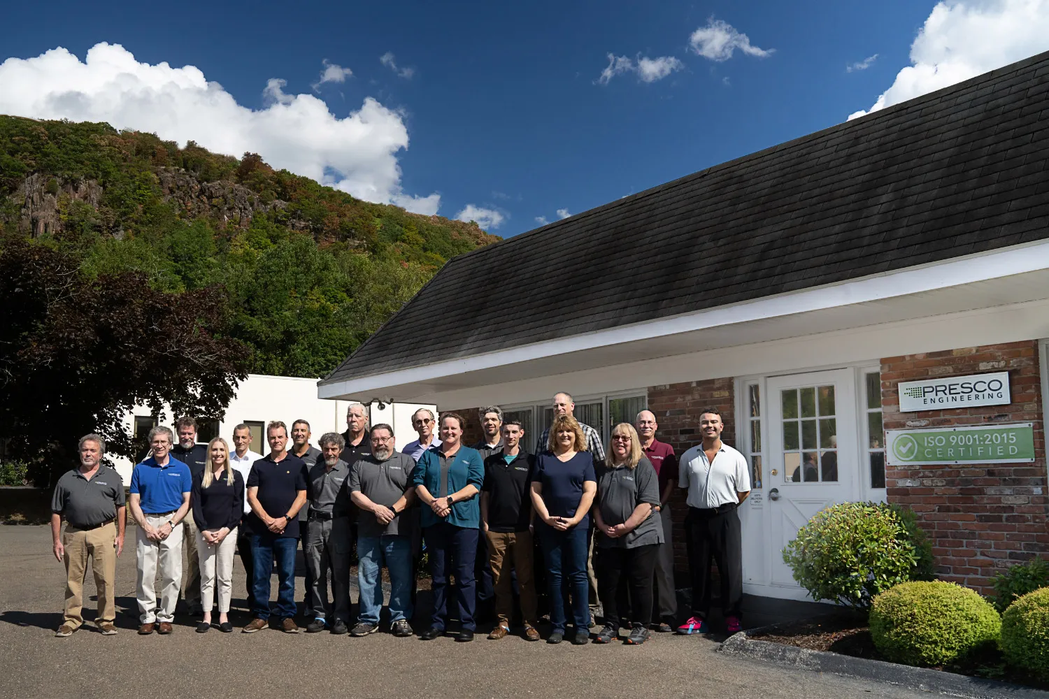 Group of 17 people standing outside Presco Engineering building under a sunny sky with trees and hills in the background.