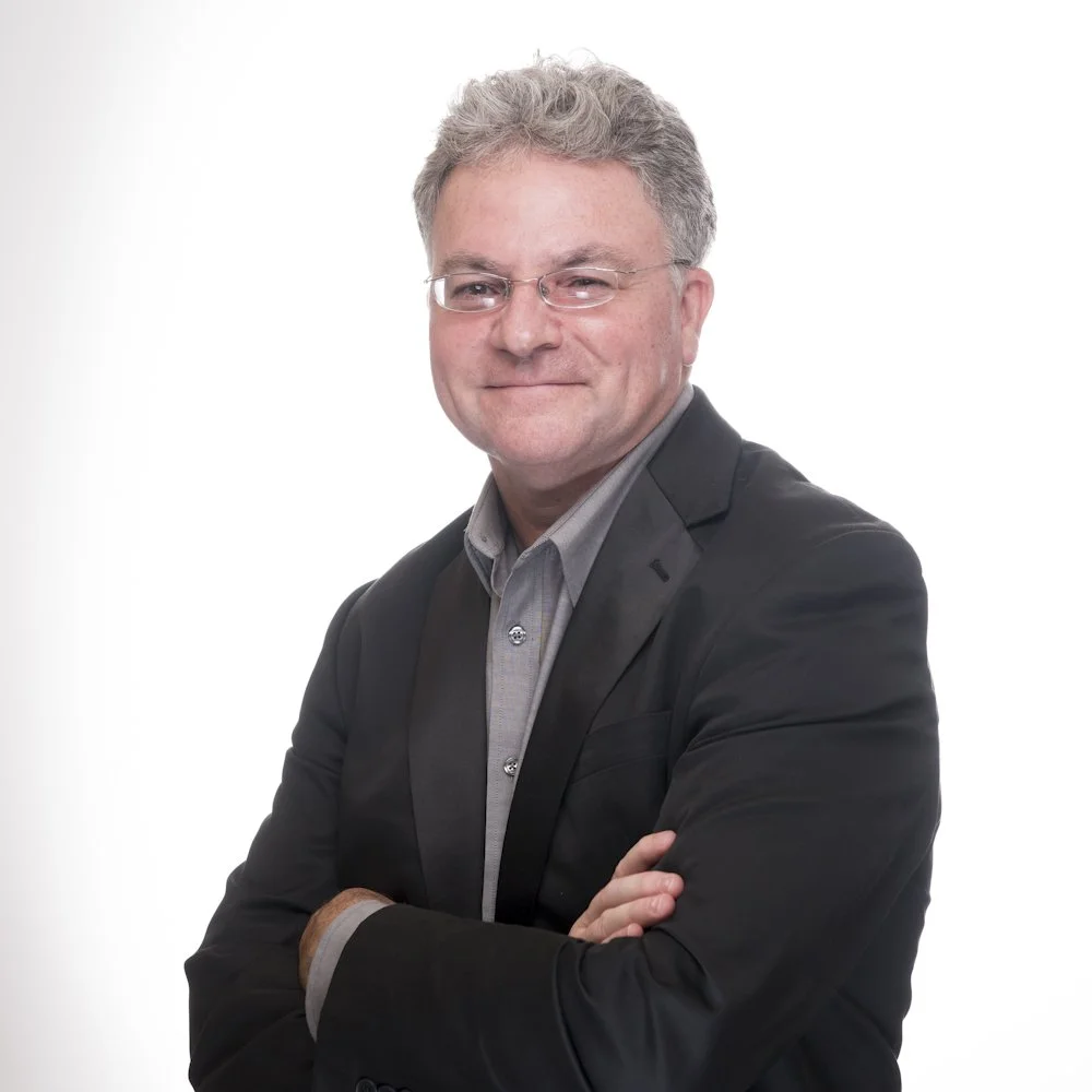 Smiling middle-aged man with gray hair and glasses wearing a black blazer and gray shirt, arms crossed.