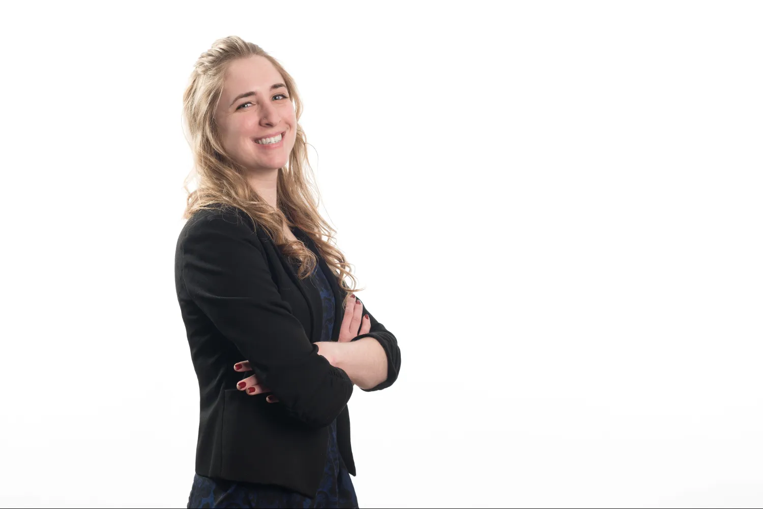 Smiling woman with long blonde hair wearing a black blazer and blue dress, standing with arms crossed against a white background.
