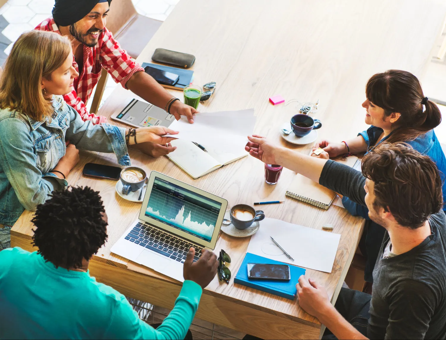 Group of five young adults collaborating around a table with laptop showing stock exchange graph, notebooks, and coffee cups.