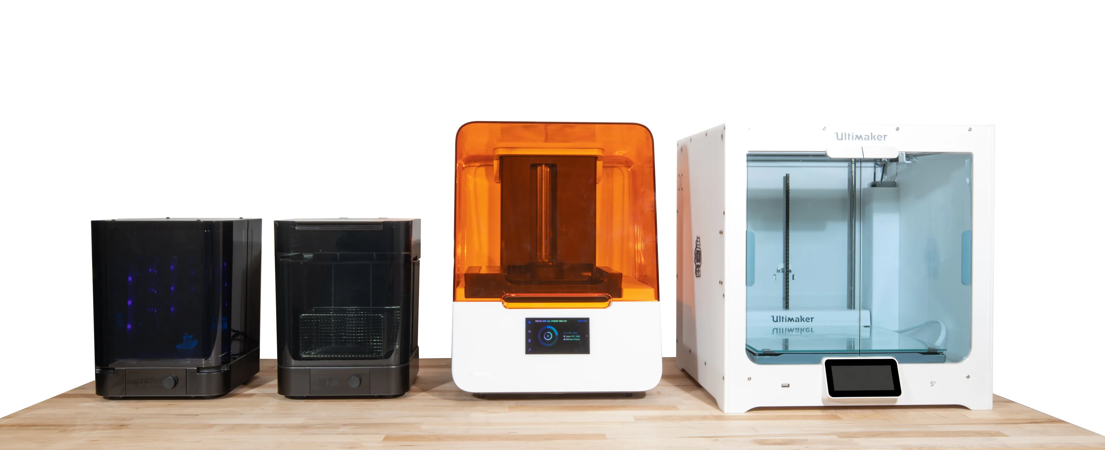 Four 3D printers of varying sizes and designs lined up on a wooden table against a white background.