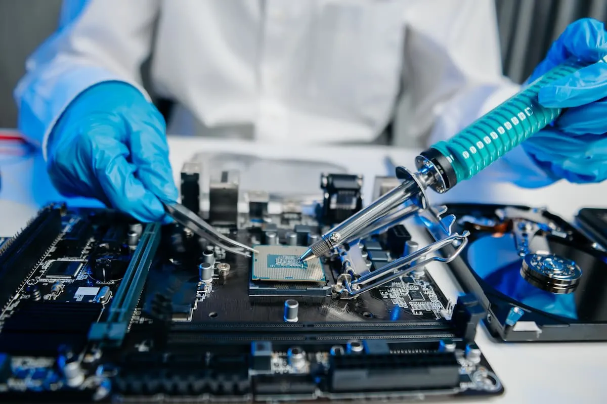 Person wearing blue gloves soldering a computer processor on a motherboard using a soldering iron and tweezers.