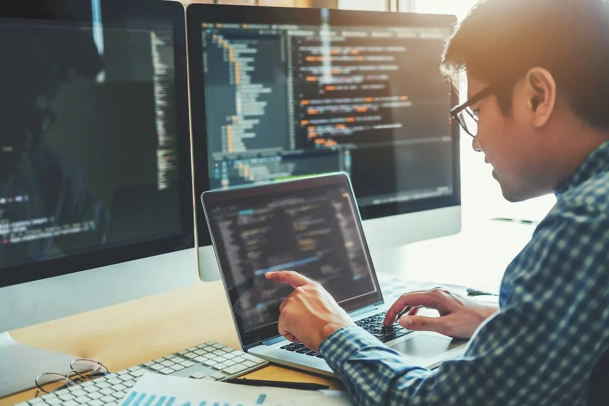 Person wearing glasses and a checkered shirt working on code on a laptop with two large monitors displaying programming code in the background.