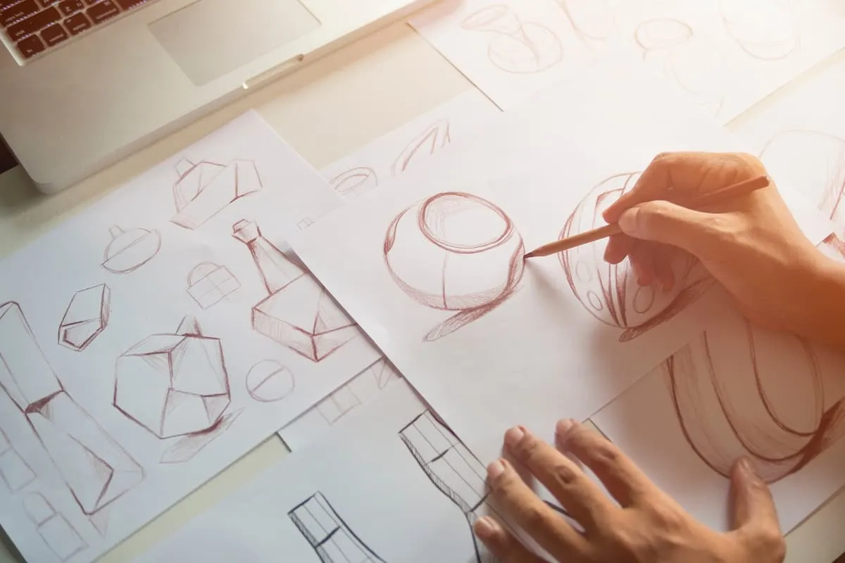 Hands sketching 3D geometric shapes and forms on white paper with a pencil next to a laptop.