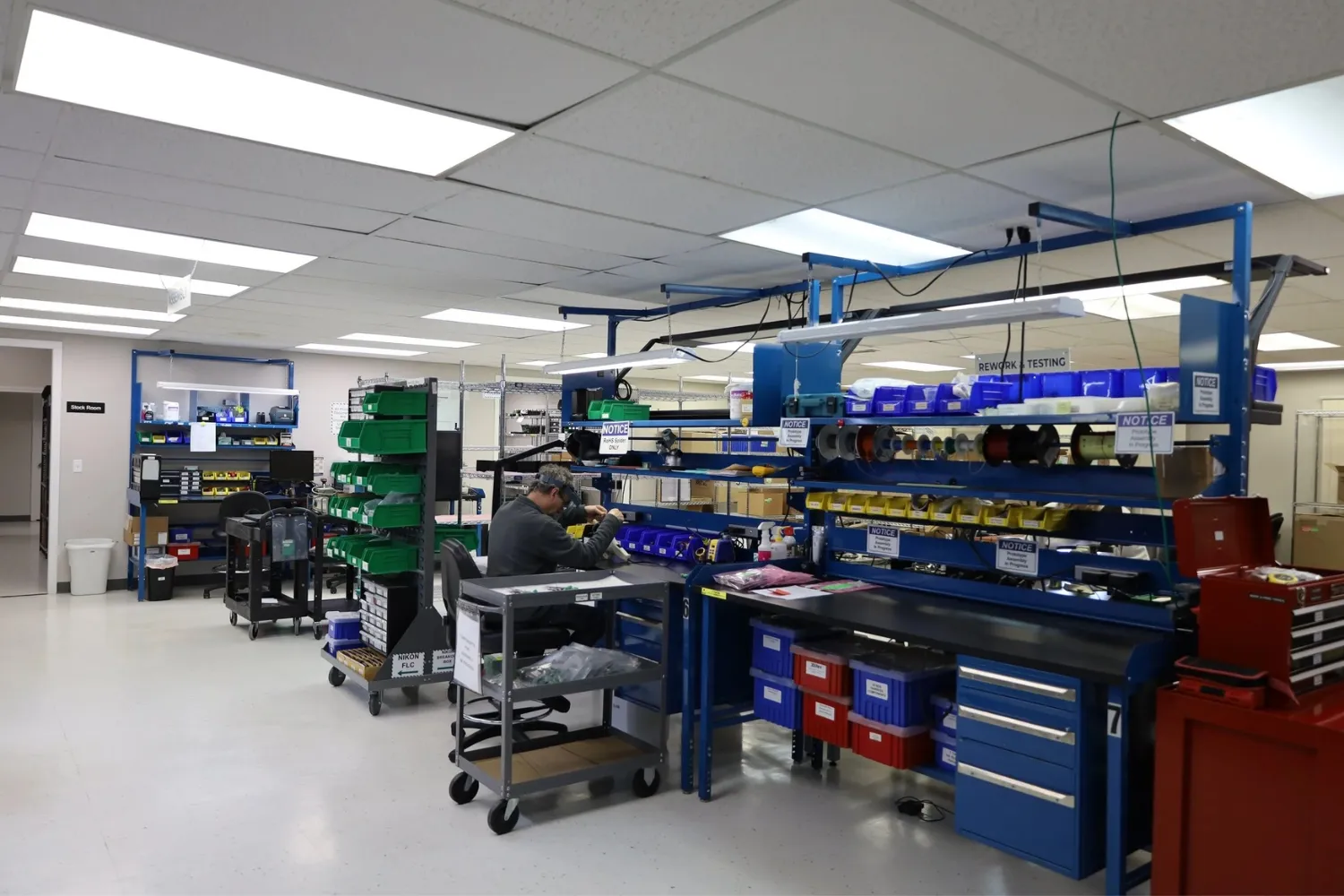 Worker sitting at an organized workstation in a bright industrial workshop with shelves of bins and equipment labeled for rework and testing.
