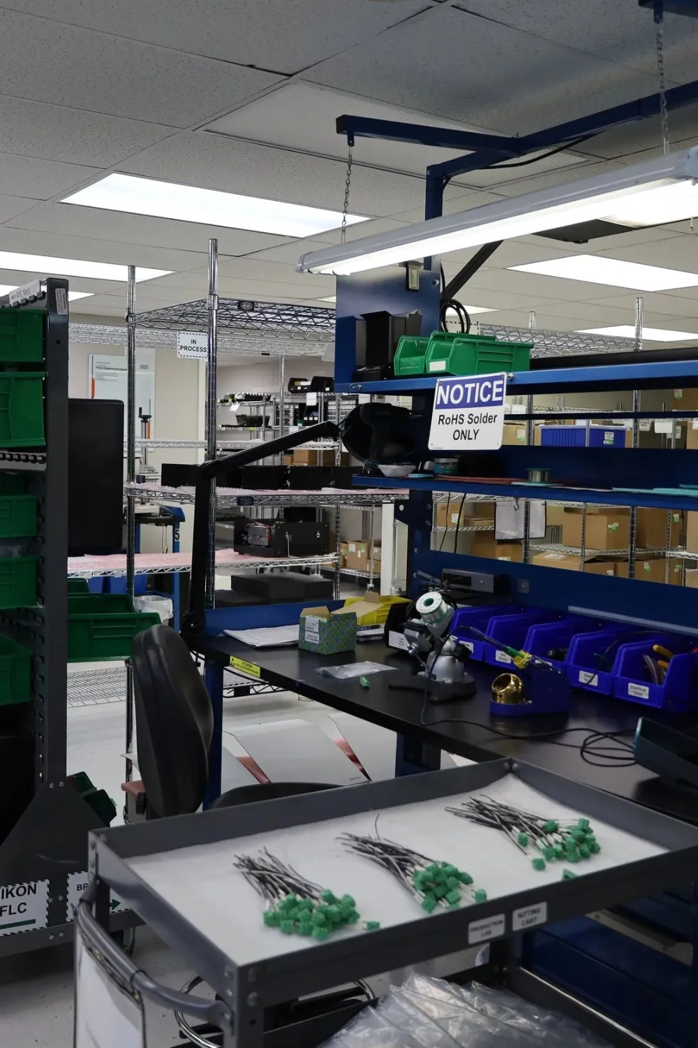 Industrial workspace with soldering station, bins for components, wires with green connectors on tray, and shelving in the background.