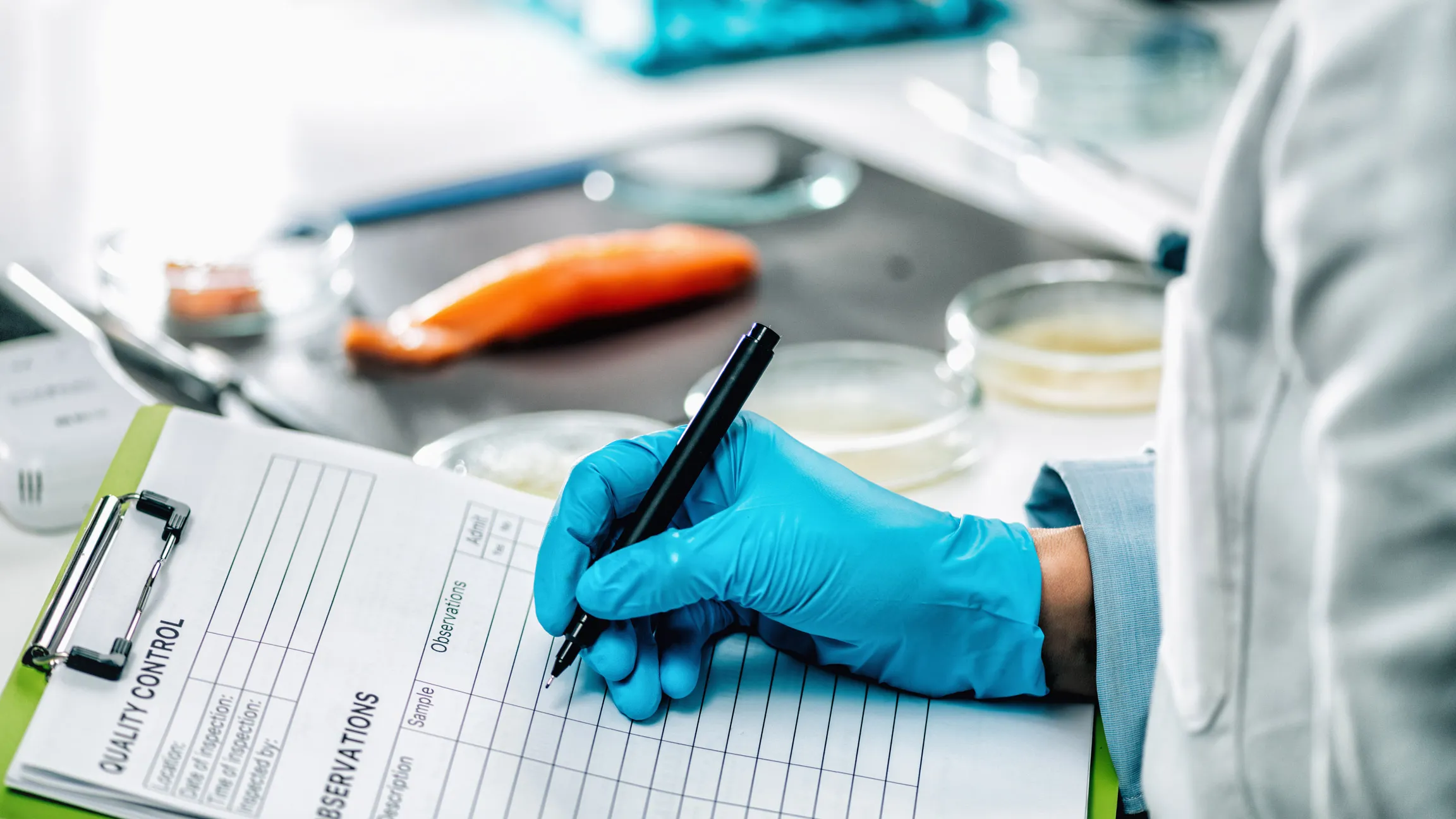 Person wearing blue gloves writing on a quality control checklist with lab equipment and a piece of salmon in the background.