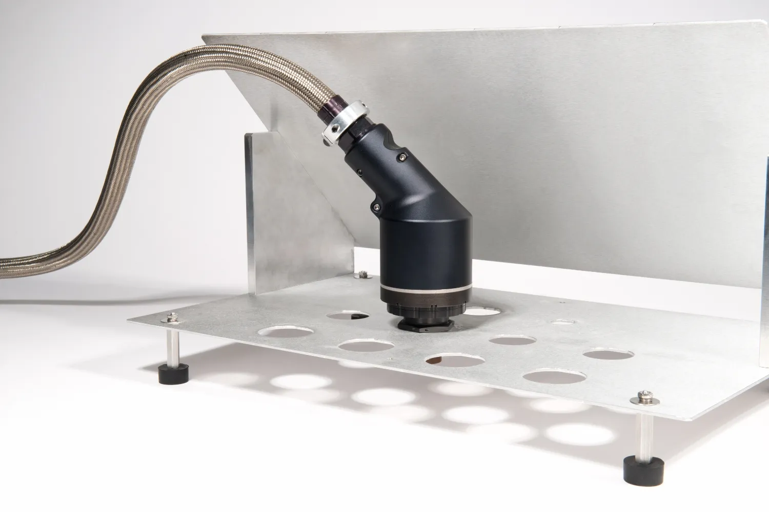 Close-up of a black industrial sensor or camera mounted on a metal plate with multiple circular holes, connected to a braided metallic cable.