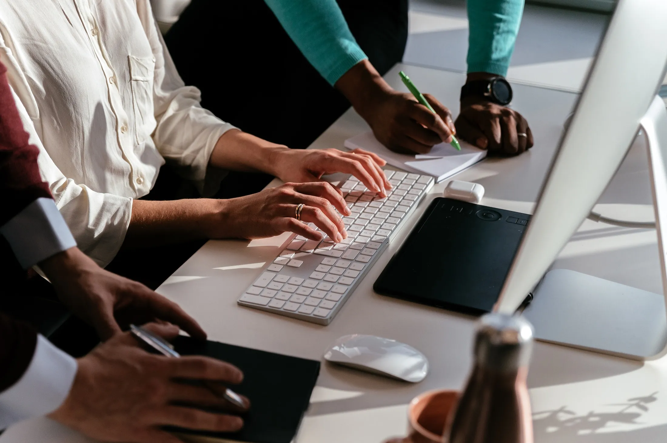 Close-up of three people collaborating at a desk with one typing on a keyboard, another writing on a notepad, and a third holding a pen and notebook.
