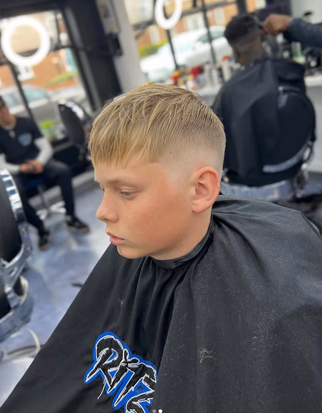 Side profile of a young boy with a fresh short blonde fade haircut sitting in a barber chair covered with a black cape.