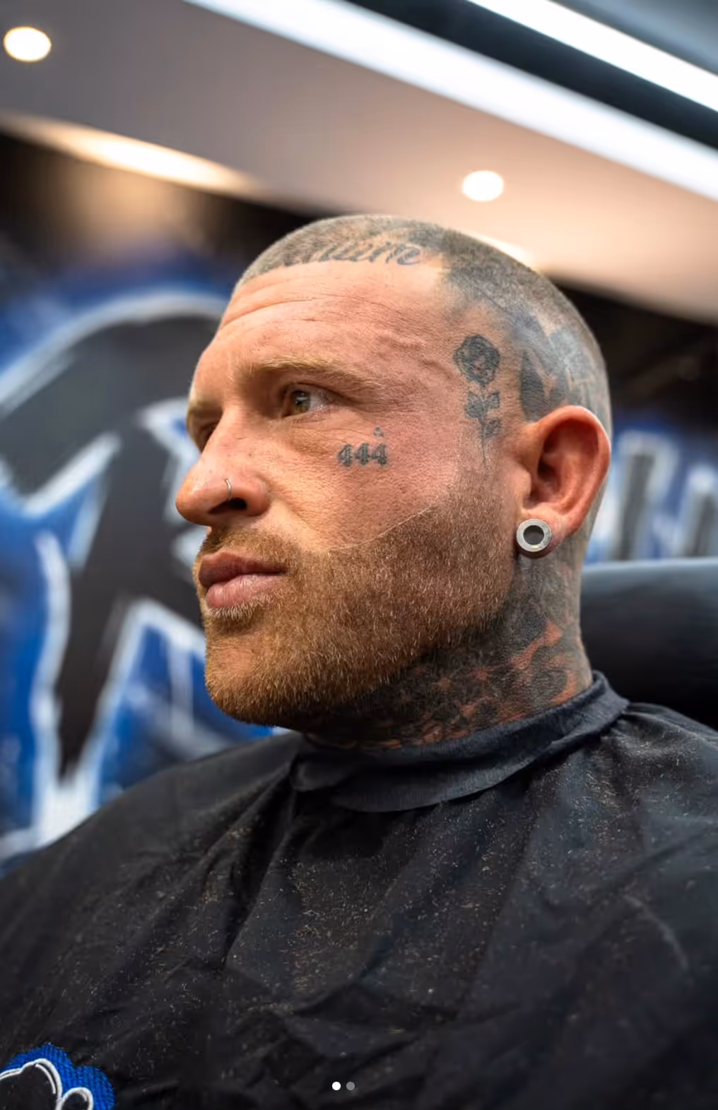 Side profile of a man with tattoos on his face and head, a nose ring, and a large ear gauge, wearing a black barber cape.