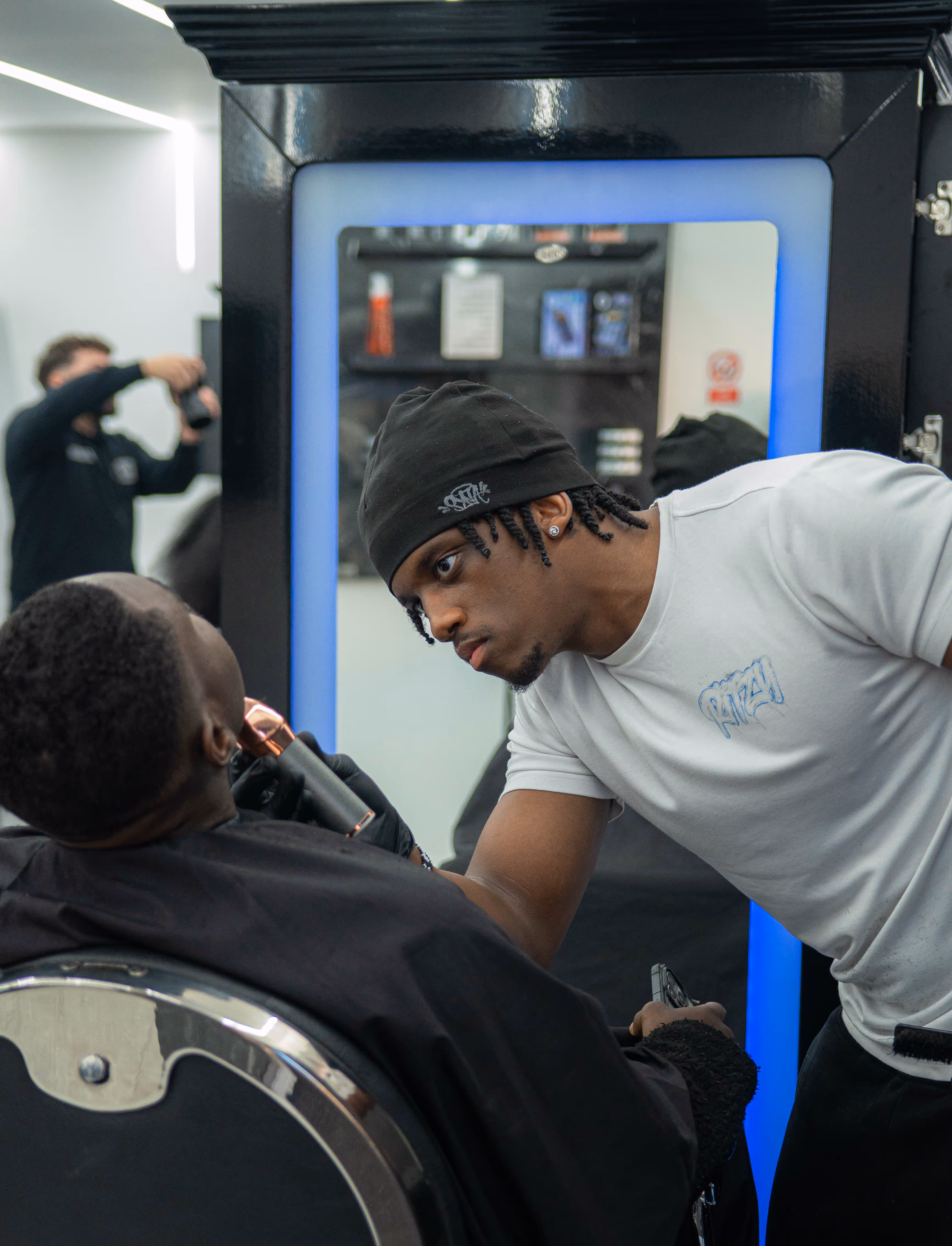 Barber wearing a black beanie intensely trimming a seated customer's beard in a modern barbershop.