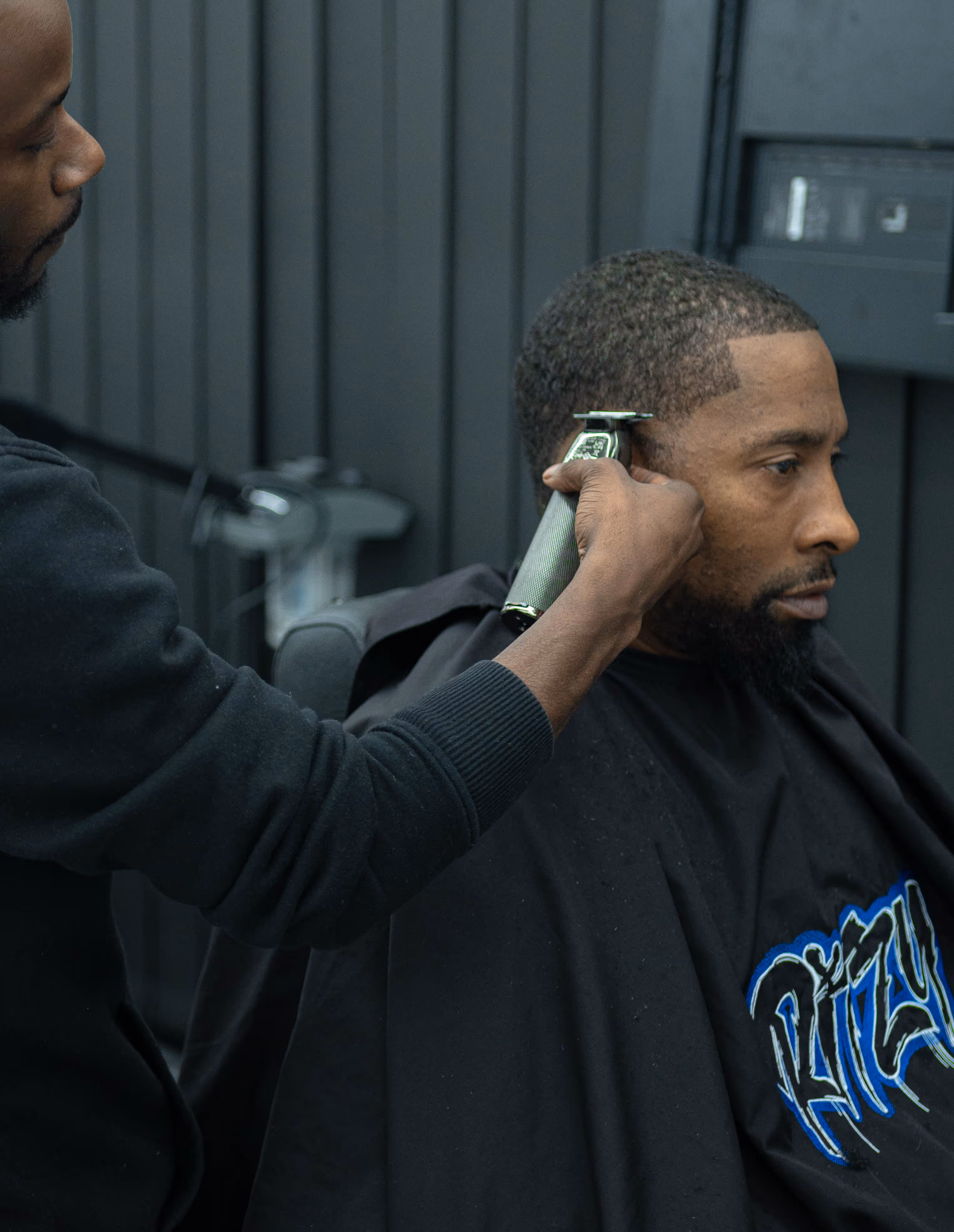 Barber using clippers to trim the hair of a seated man with a beard and short haircut.
