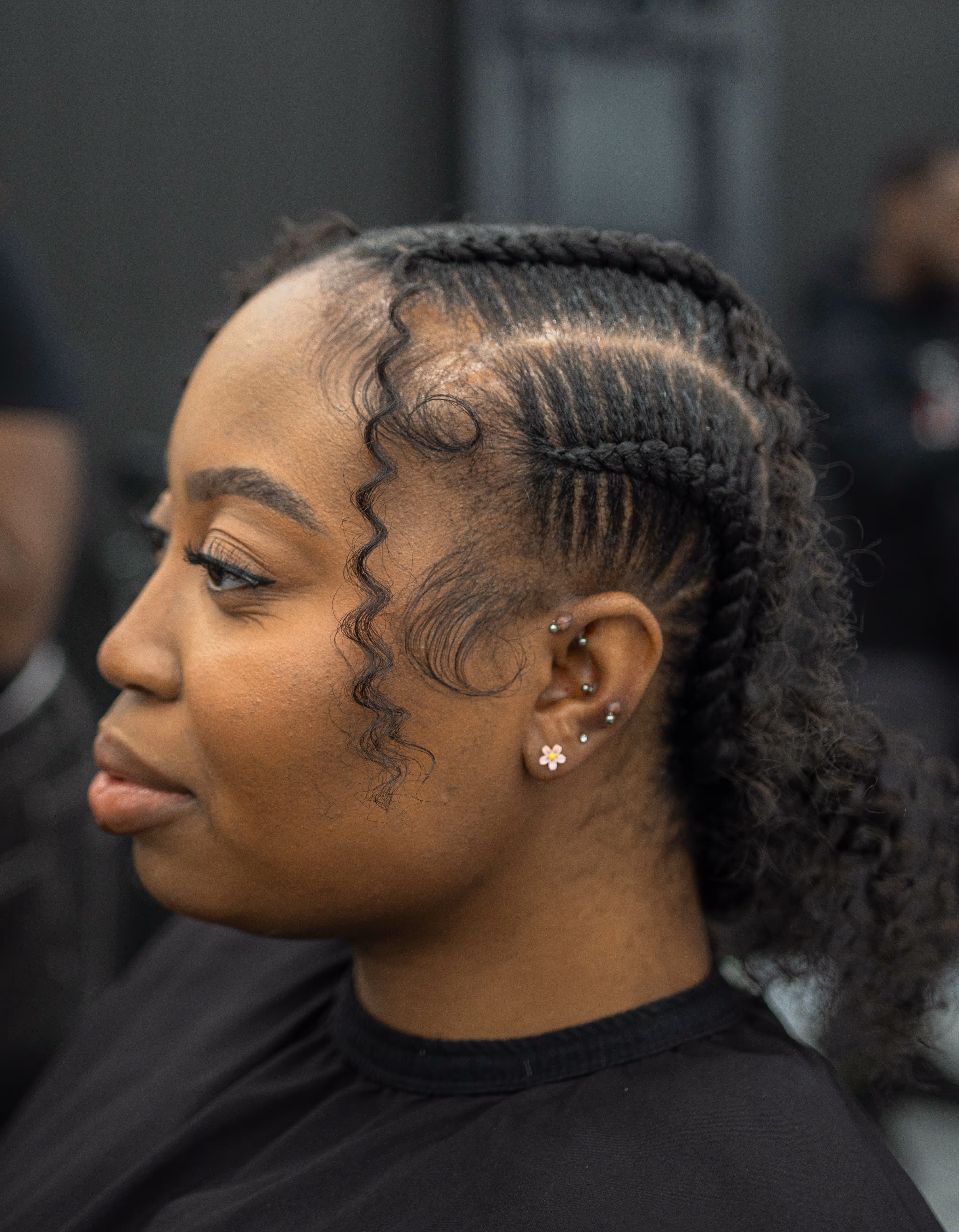 Profile of a woman with neatly braided hair styled in cornrows and multiple ear piercings, including a floral stud earring.