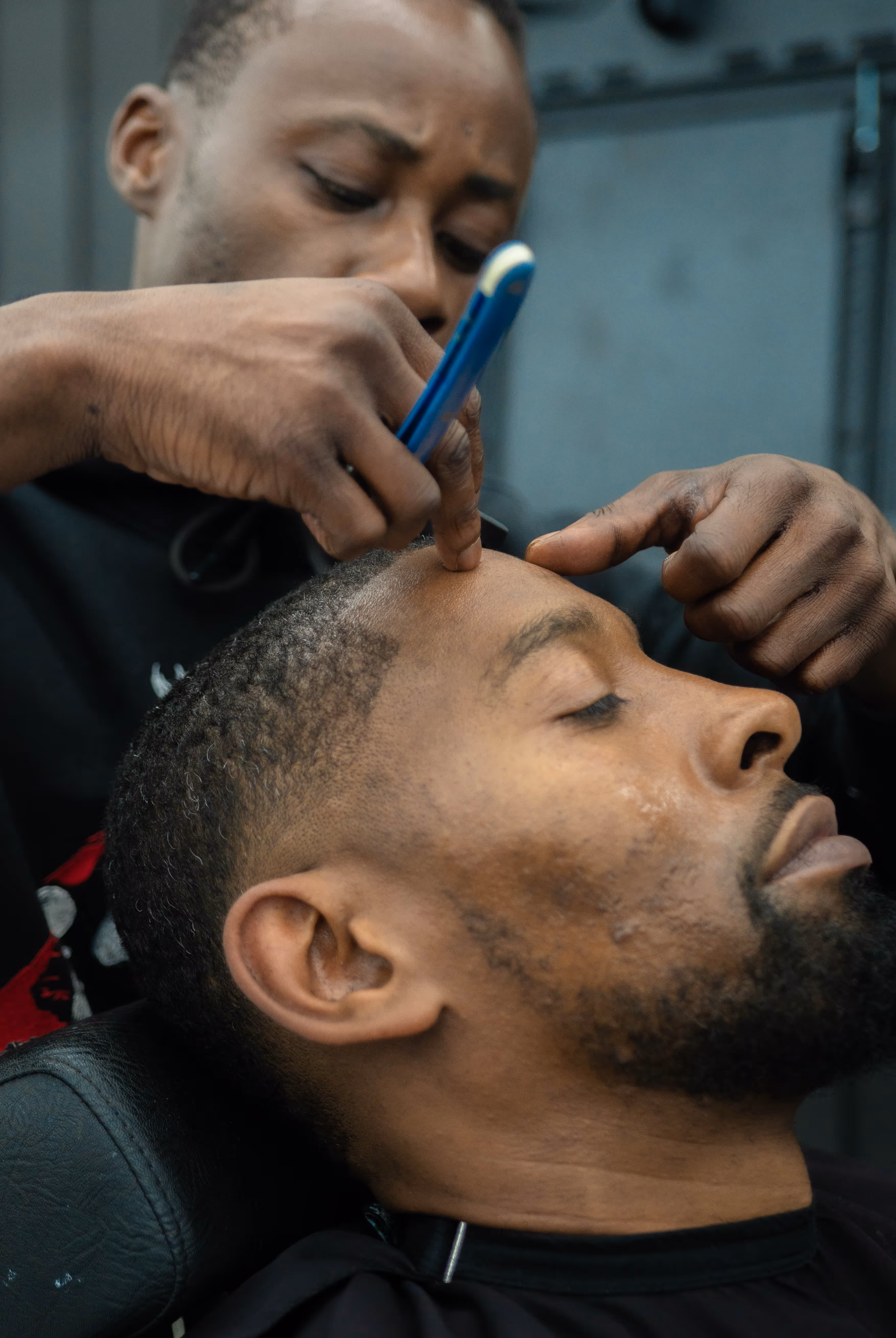 Barber using a straight razor to shape the hairline of a seated man with eyes closed.